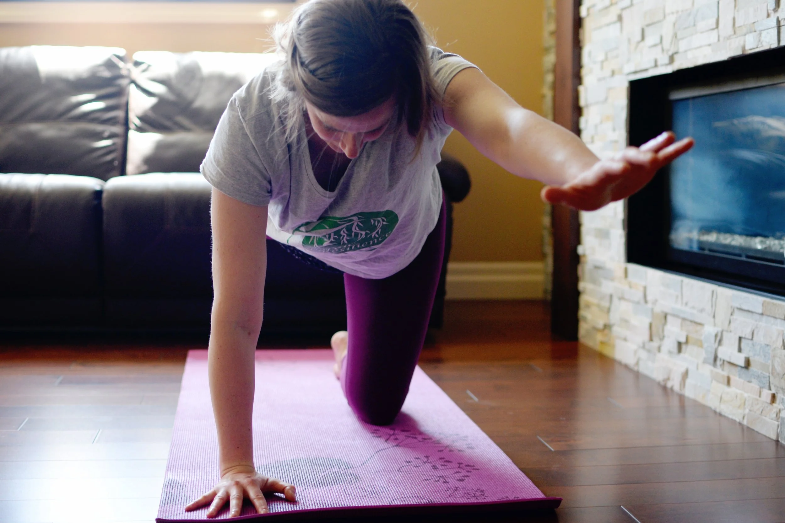Lara Desrosiers of Pelvic Resilience doing yoga on a pink mat indoors, reaching toward the floor with her left hand, with a couch and a stone fireplace in the background.