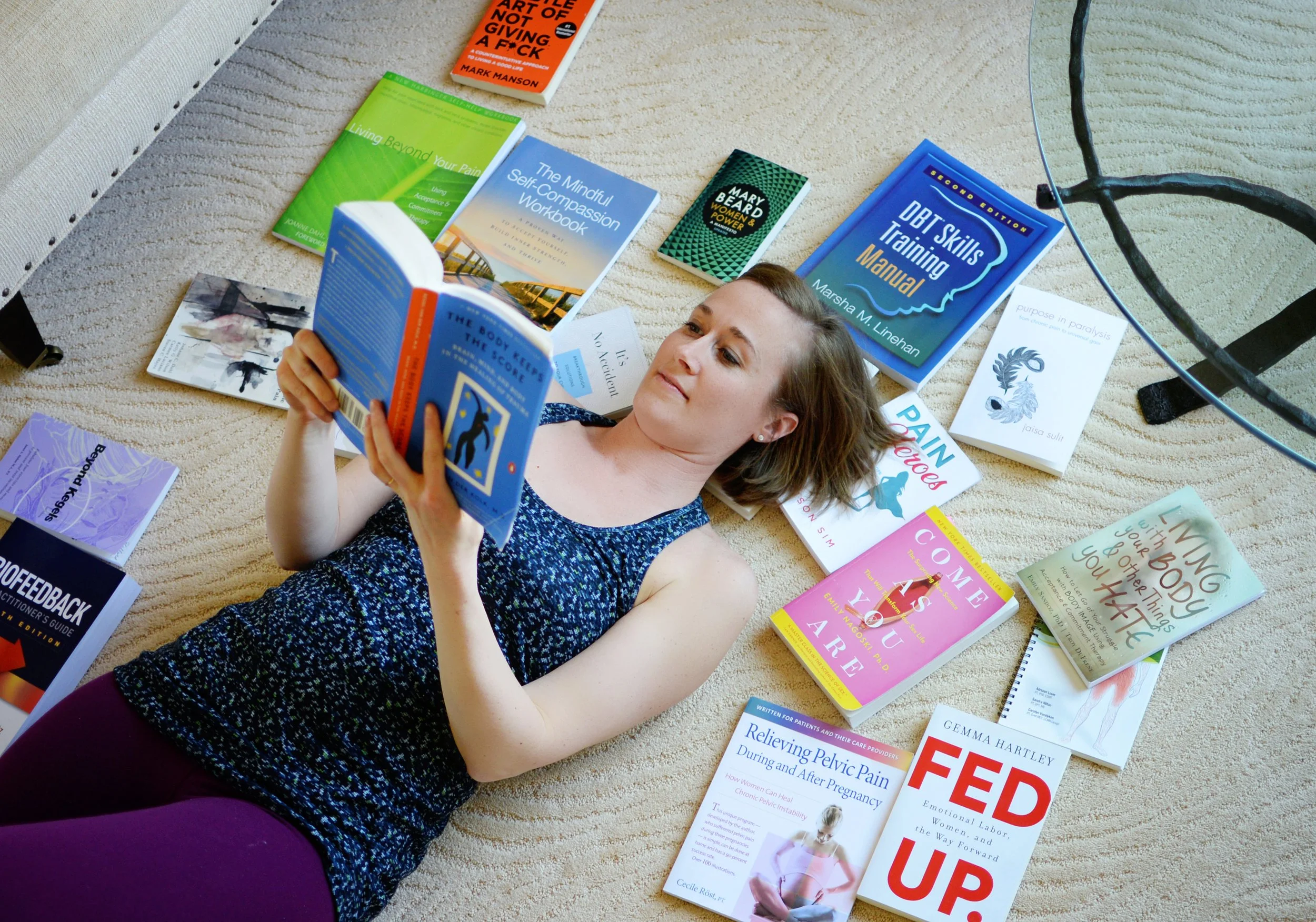 Lara Desrosiers of Pelvic Resilience lying on a beige carpet surrounded by books about health, self-help, and personal development, reading a blue book titled 'The Body Keeps the Score'.