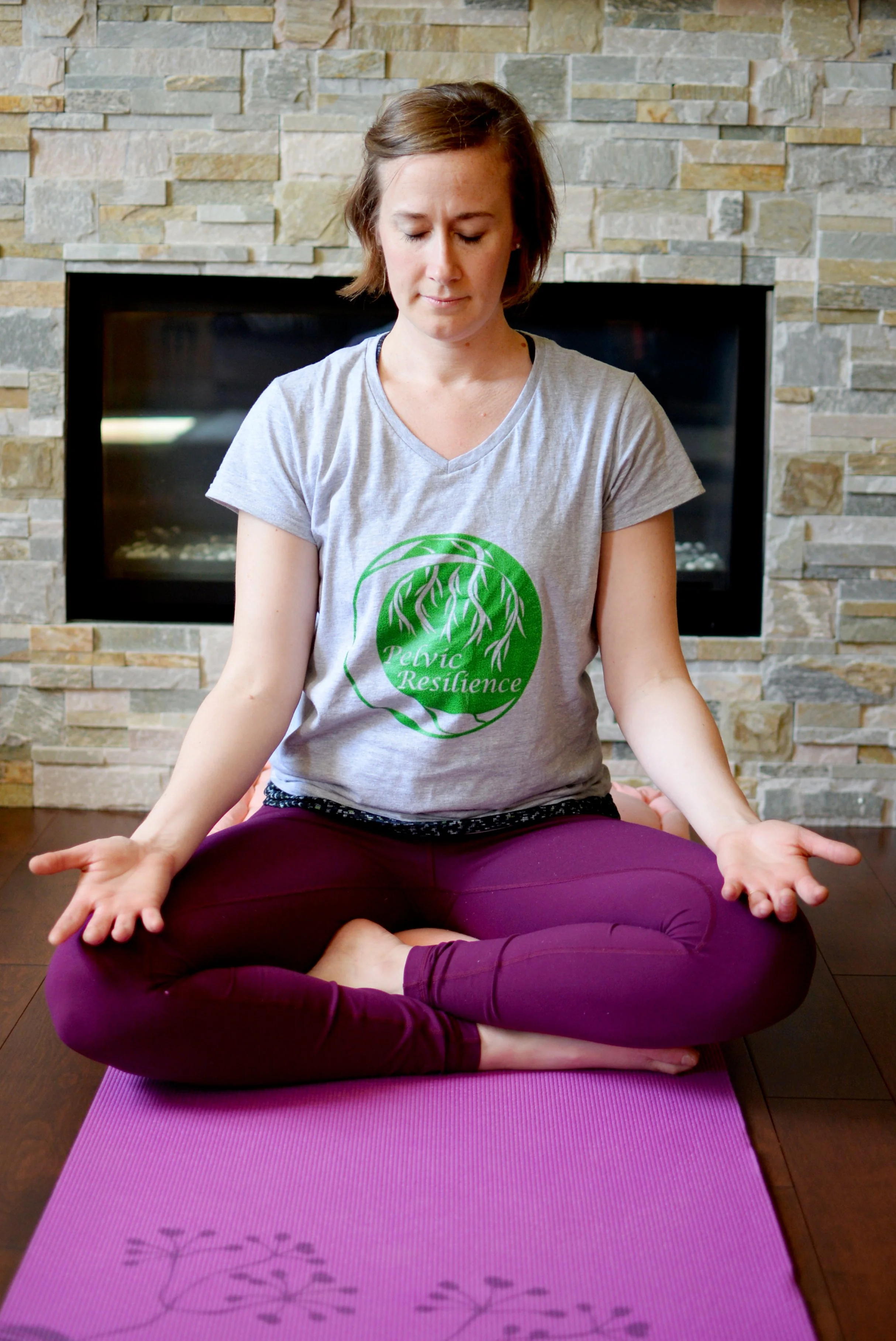 Lara Desrosiers of Pelvic Resilience performing a seated meditation or yoga pose on a purple mat in front of a stone fireplace, wearing a gray t-shirt with a green logo that reads 'Pelvic Resilience' and purple leggings.