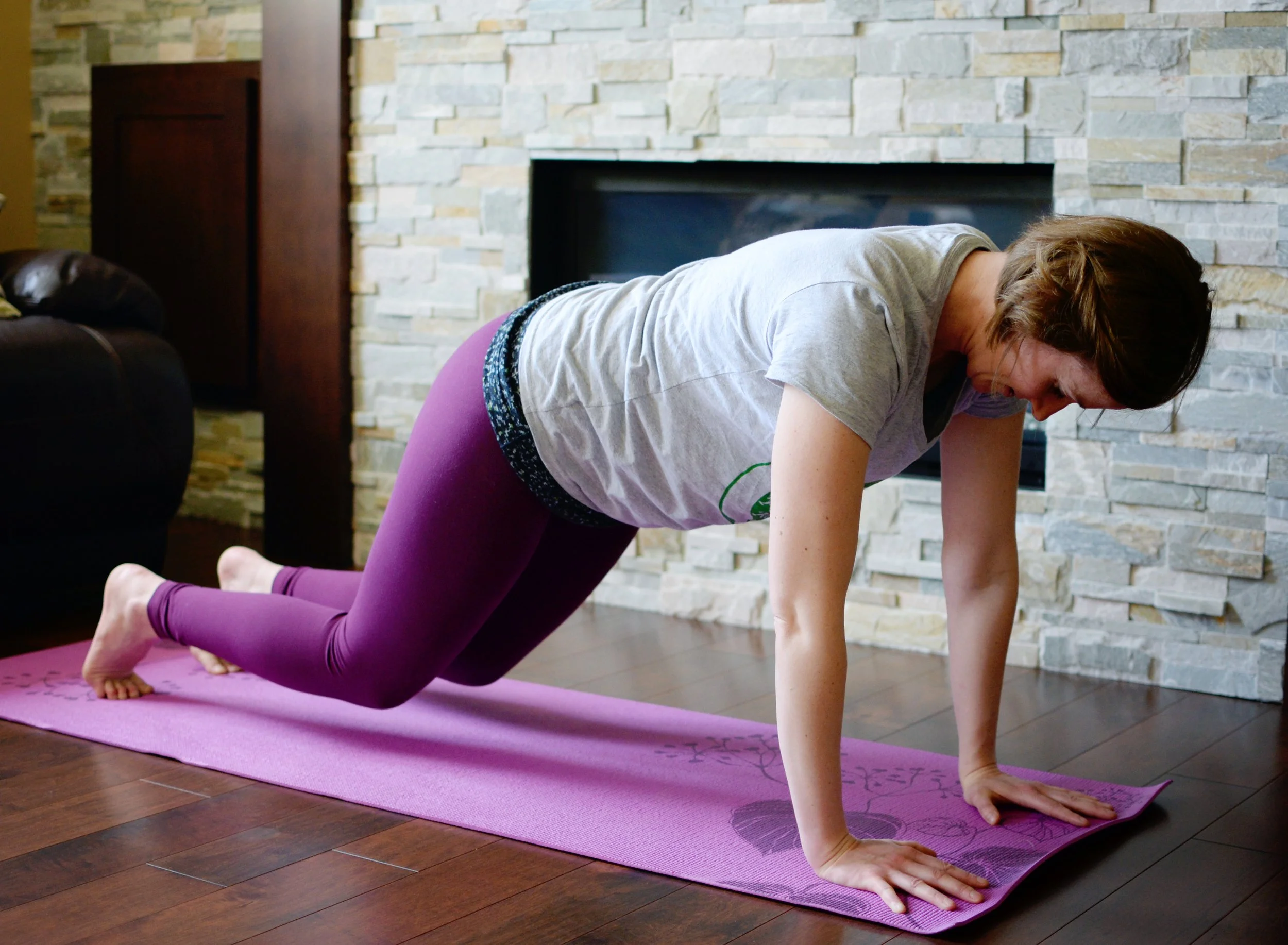 A woman practicing yoga on a pink mat in a living room with a stone fireplace, using a tabletop pose.