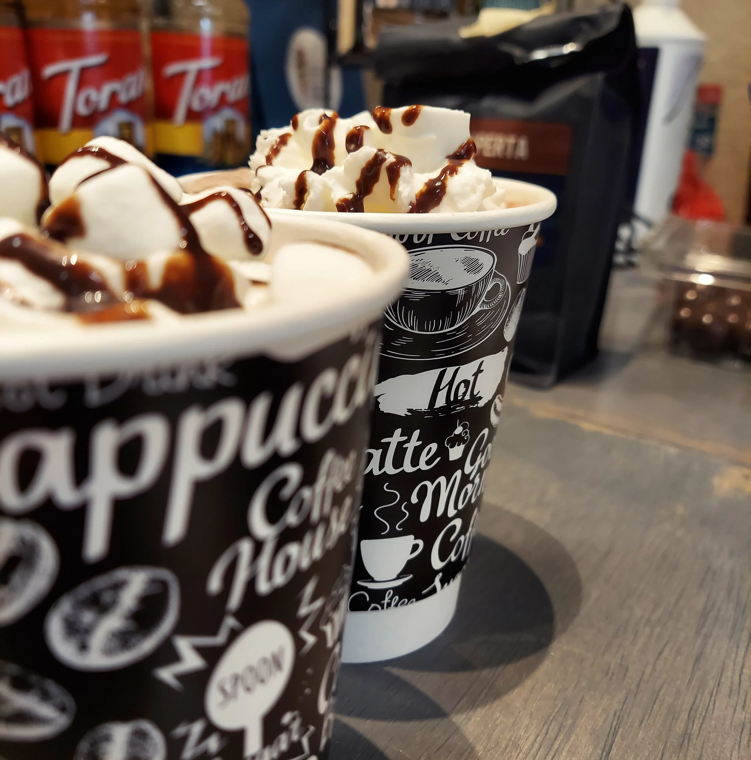 Two coffee cups topped with whipped cream and drizzled with chocolate sauce, on a wooden table in a café setting.