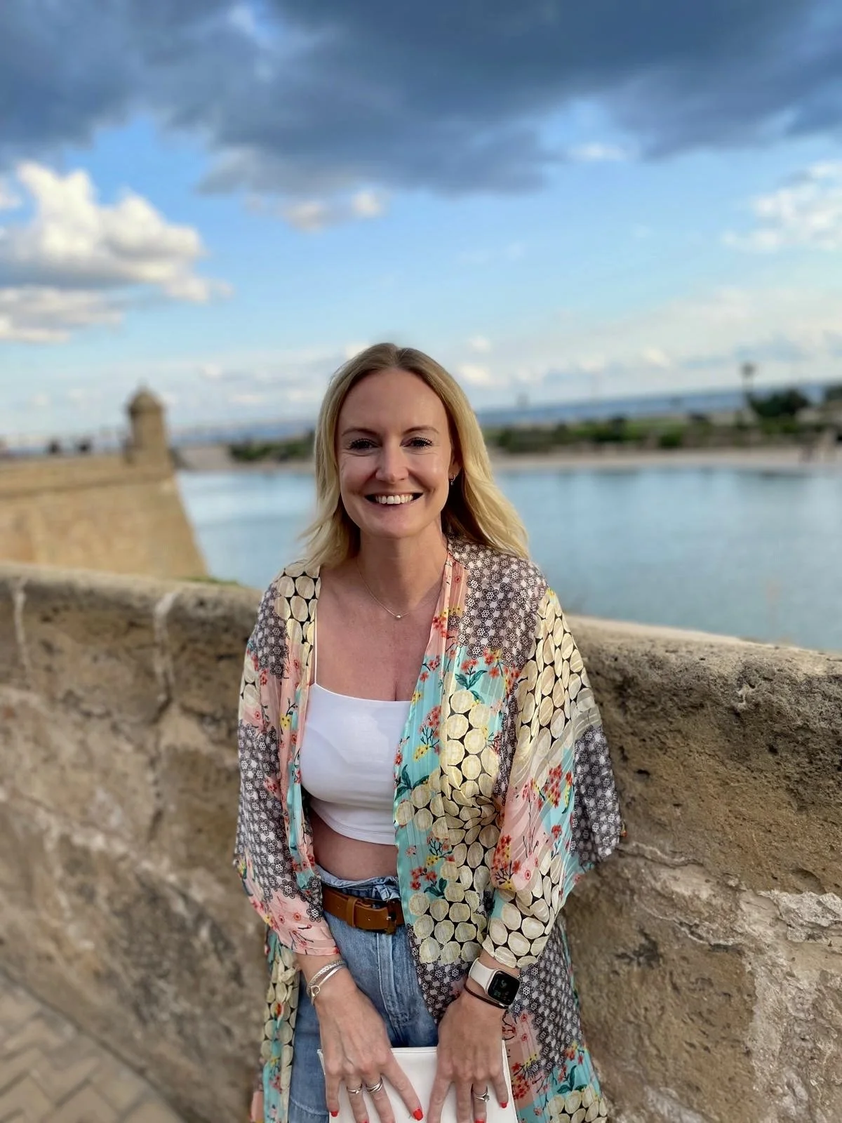 A woman with blonde hair smiling, standing outdoors by a stone wall near water, with a cityscape and cloudy sky in the background.