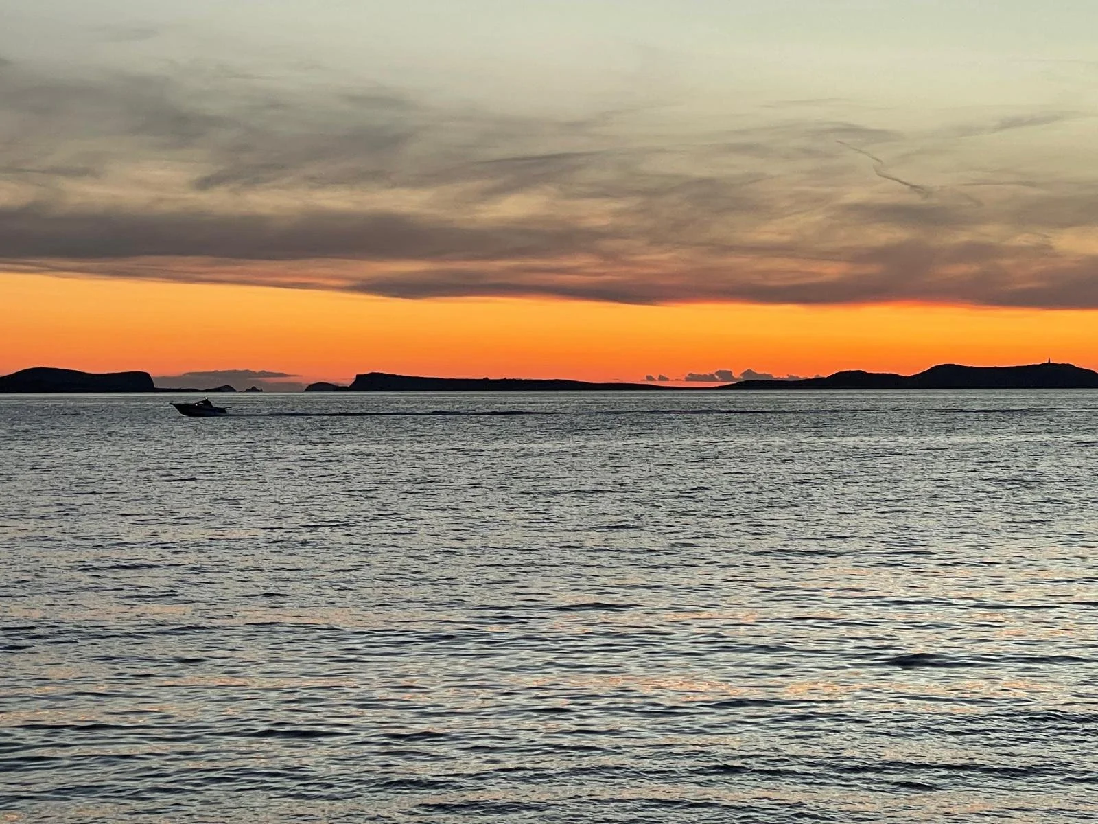 A boat moving across the water at sunset, with silhouetted islands and a vibrant orange and gray sky.