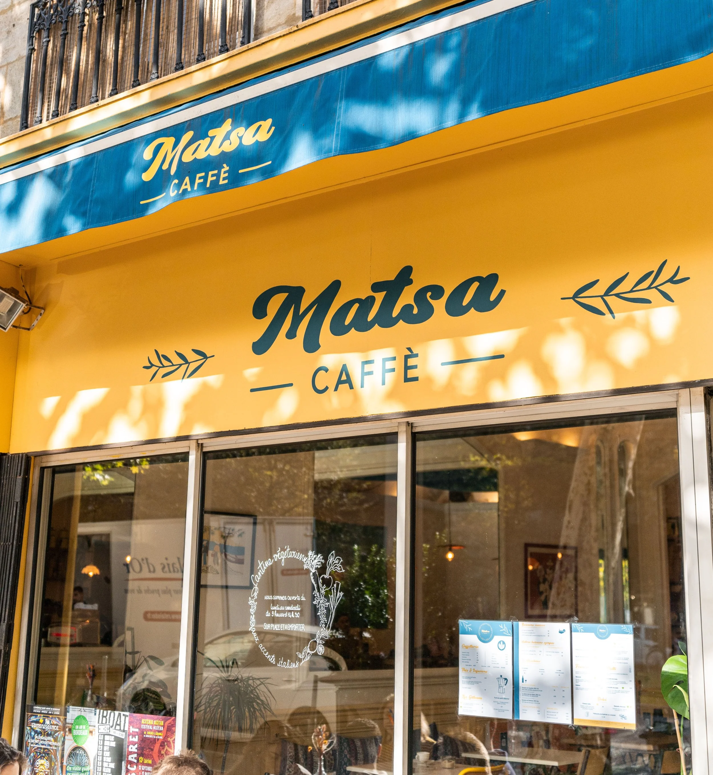 Facade du café 'Matsa Caffè' avec un panneau jaune et un auvent bleu, vitres avec affiches et menu, intérieur visible avec clients.