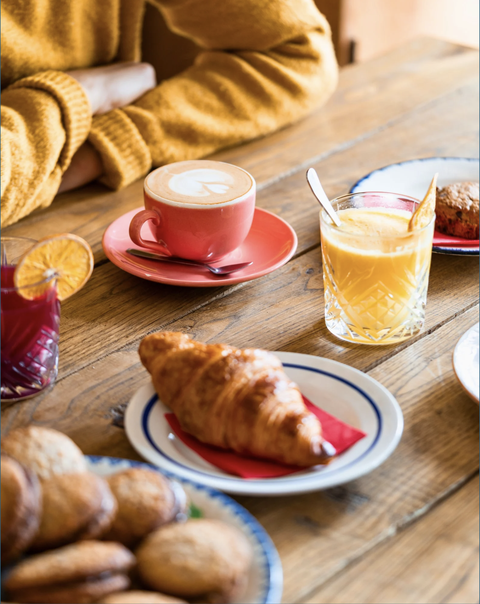 Petits-déjeuners avec café, orange pressée, croissant, muffin, et boissons colorées sur une table en bois.