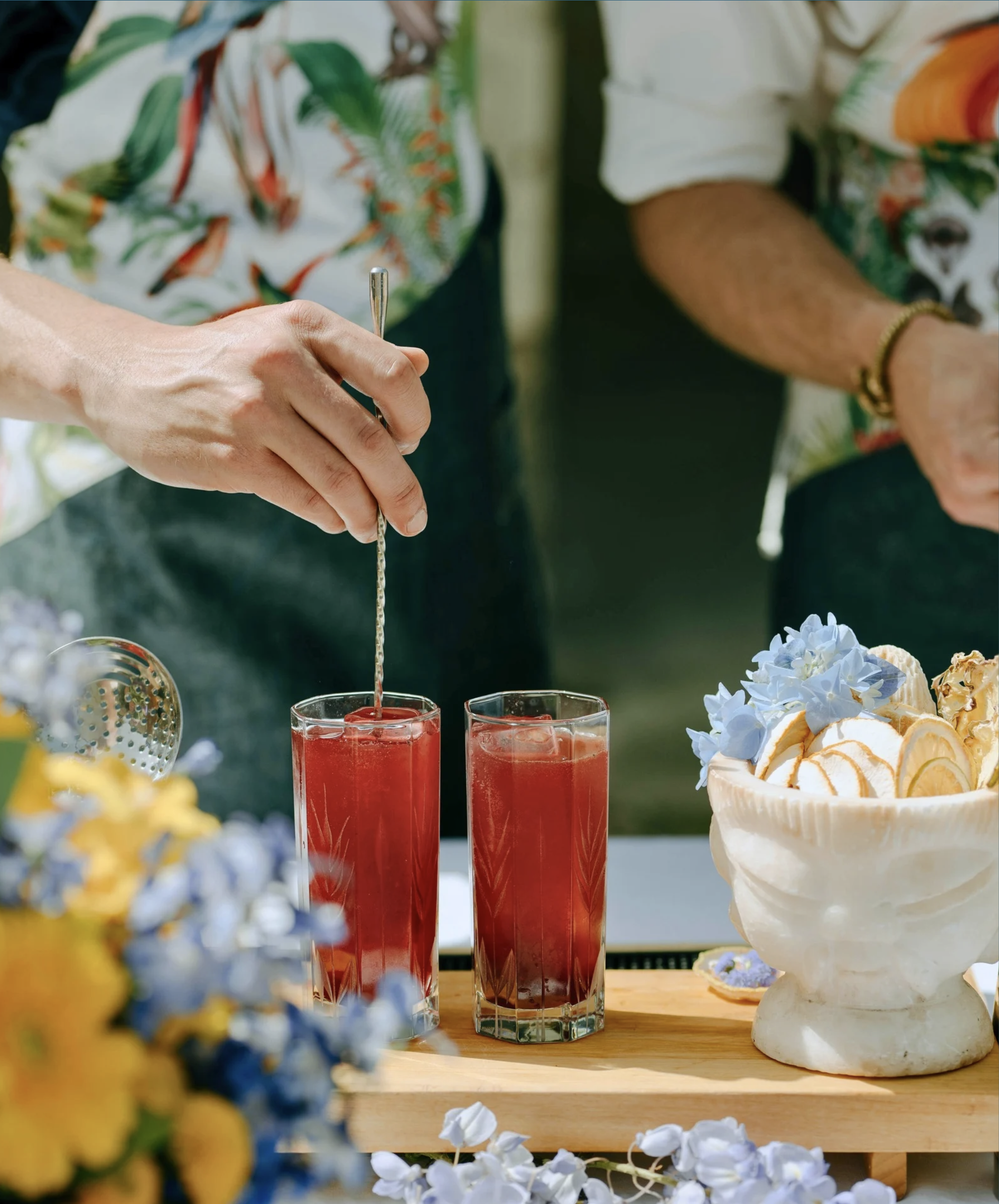 Deux personnes préparent des cocktails rouges dans des verres avec des glaçons. Il y a un bol de glaces ou de sorbets avec des morceaux de citron et des fleurs, et des fleurs jaunes et bleues en premier plan.