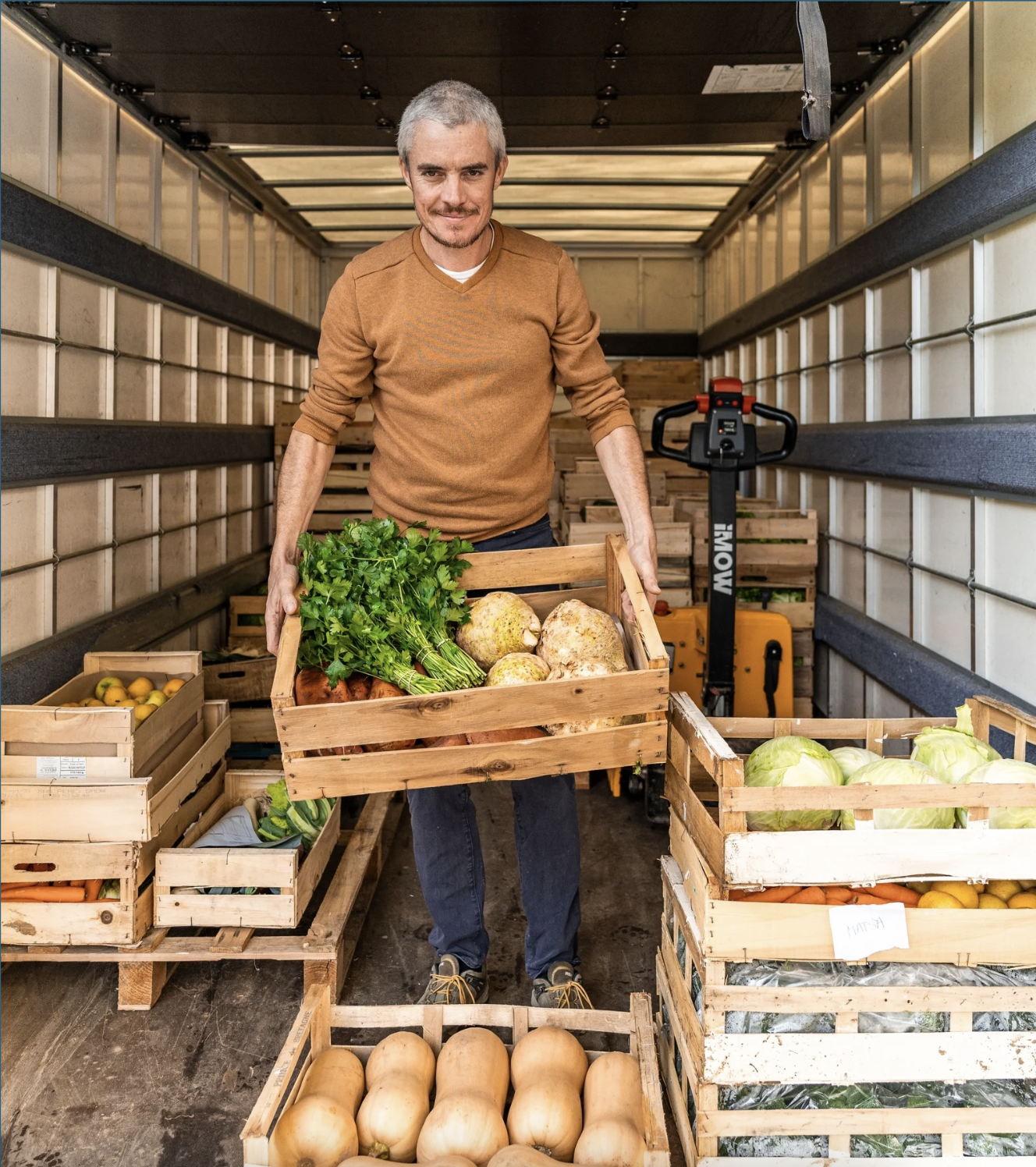 Homme dans un camion de livraison de légumes, tenant une caisse de légumes verts et de racines, entouré d'autres caisses de légumes.