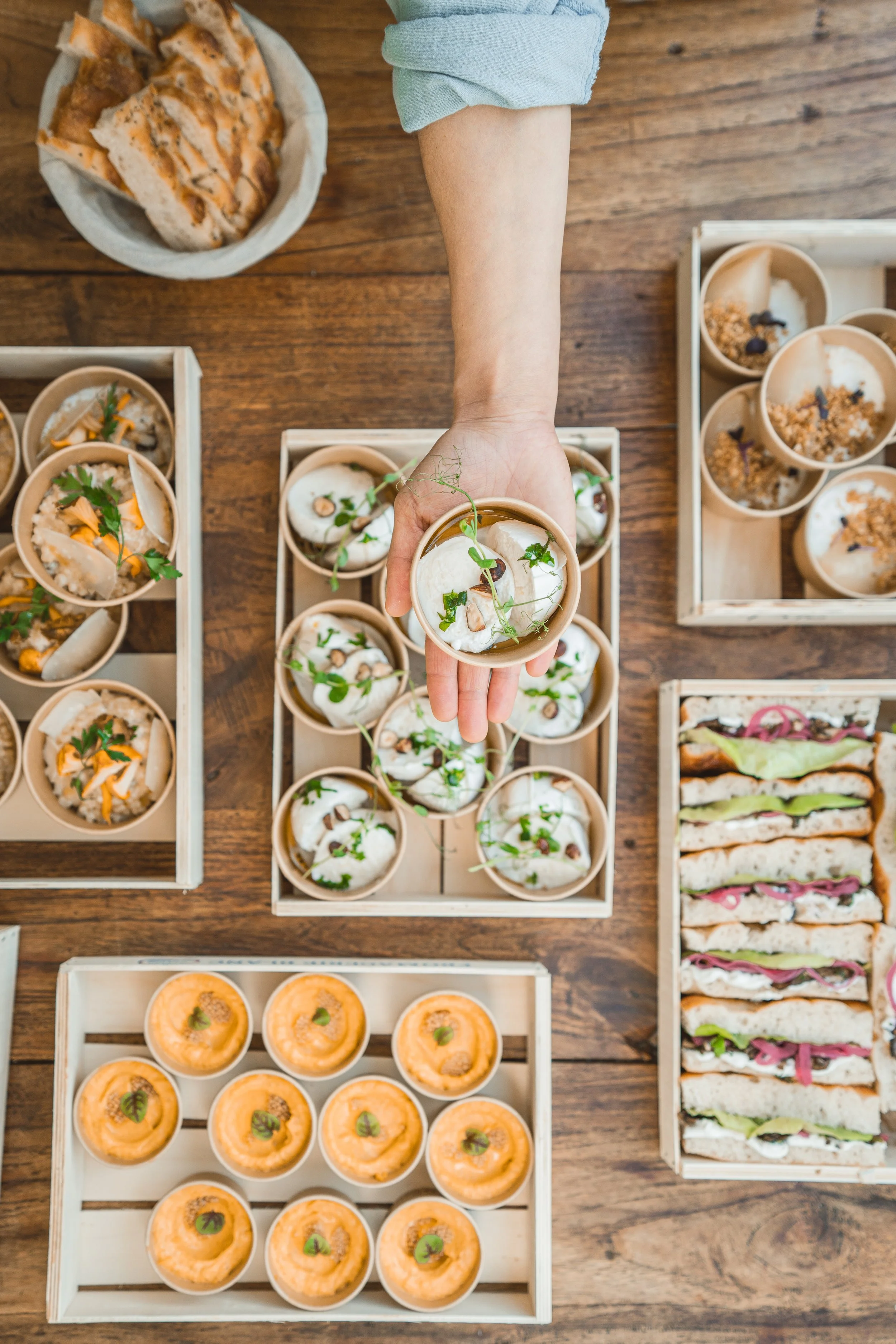 Une main tenant une coupe de soupe ou de salad, entourée de plusieurs autres coupes avec des plats variés, sur une table en bois.