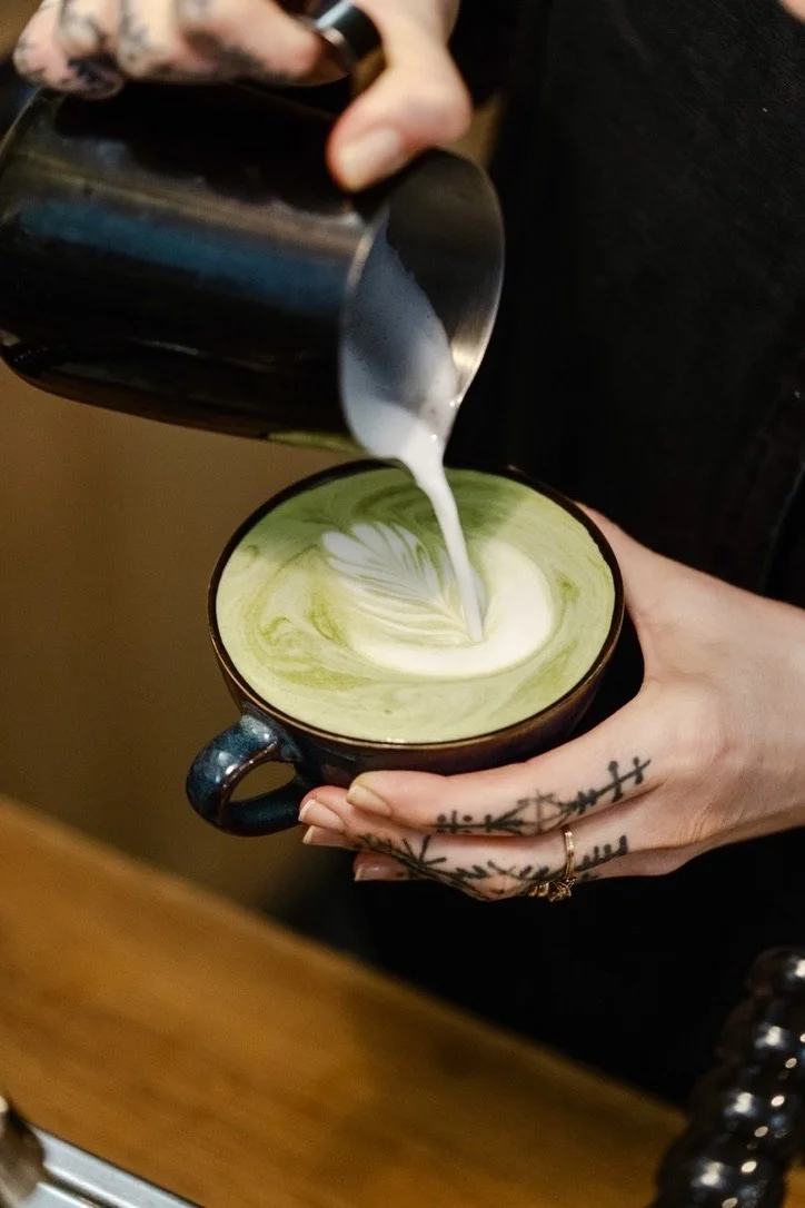 Une personne verse du lait dans un bol de matcha pour faire un latte artistique, avec des tatouages sur les doigts.