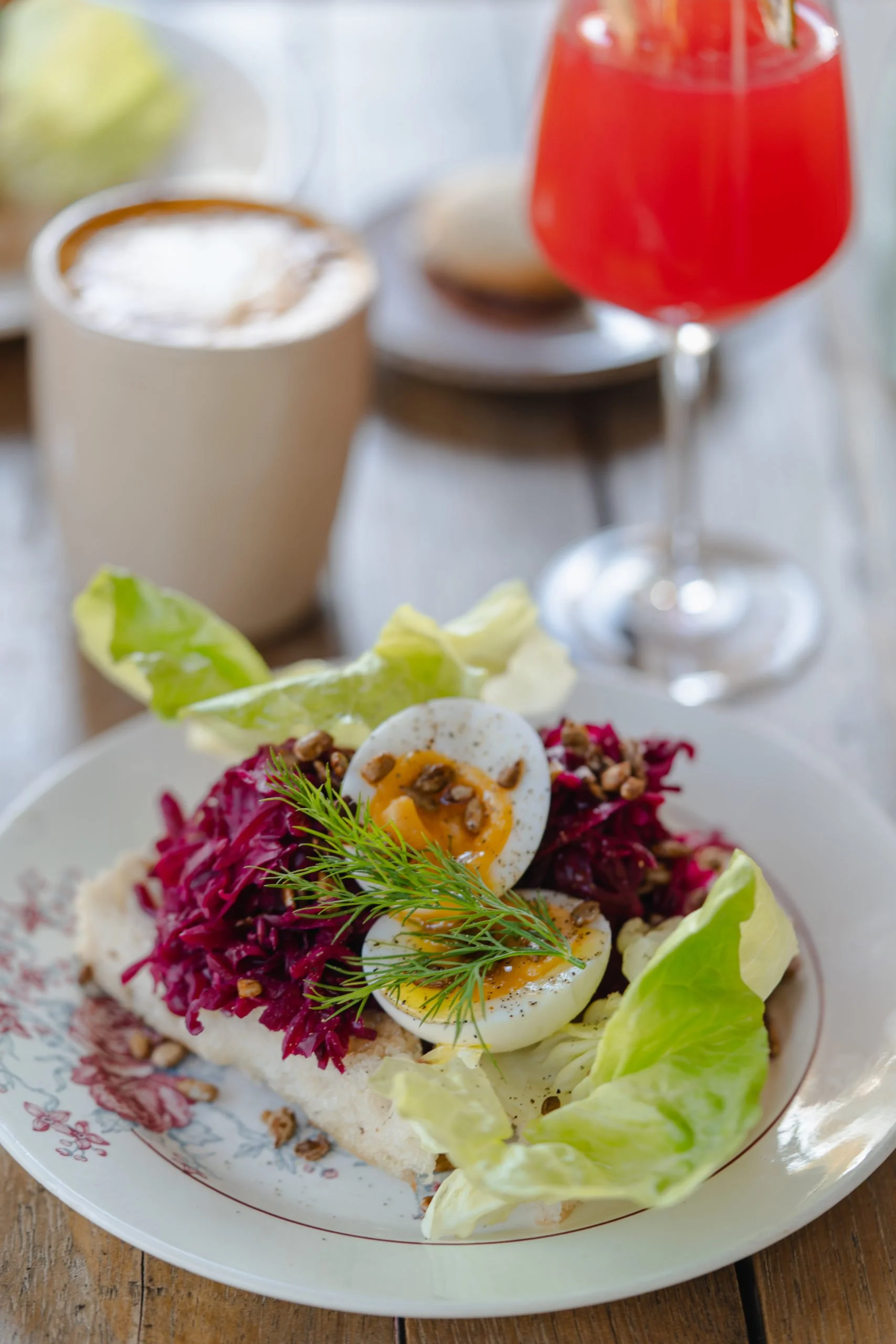 Salade avec œufs mollets, chou rouge, laitue, noix, herbes, sur une assiette, accompagnée d'une boisson rouge et d'une autre chaude, sur une table en bois.