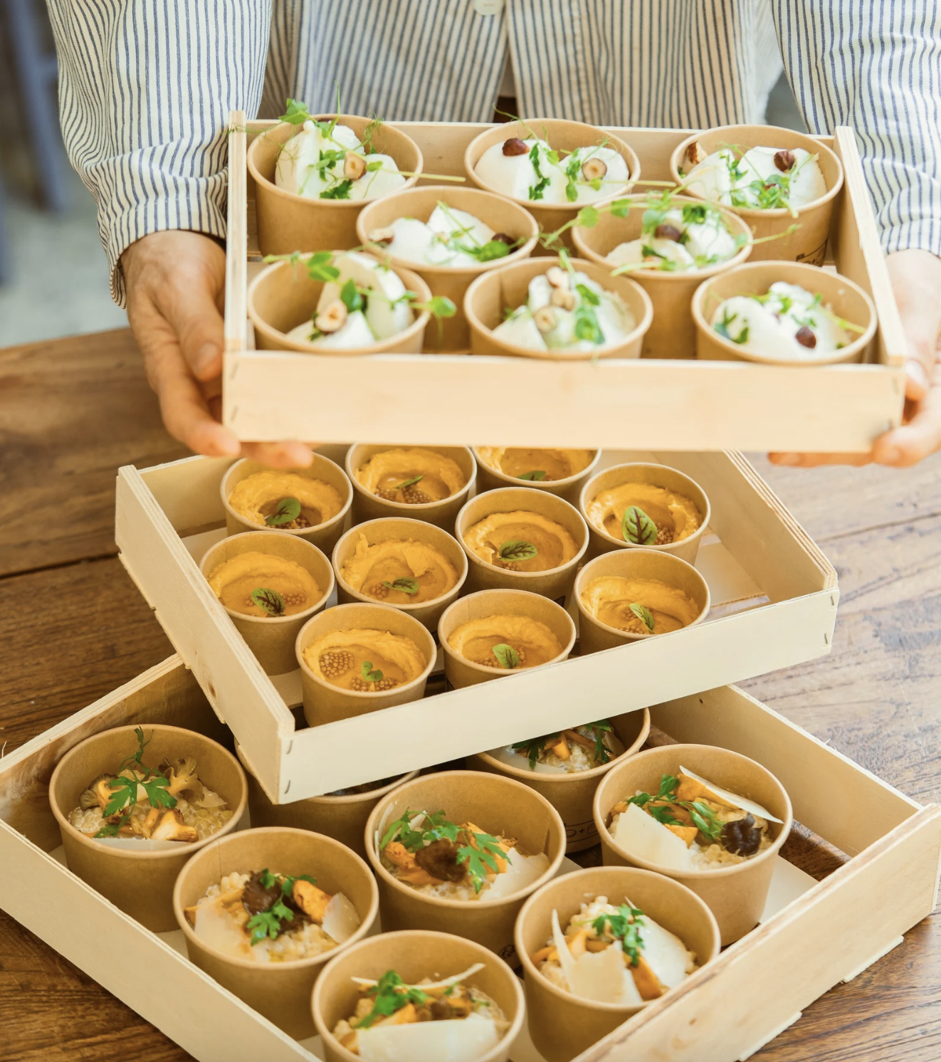 Une personne présente trois plateaux en bois contenant des petits bols de différents plats, garnis de garnitures vertes et décoratives.