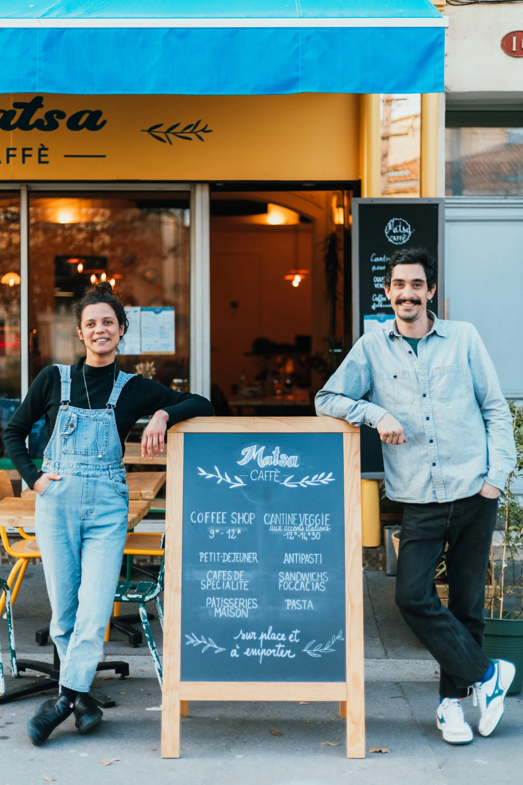 Mathieu et Sara posent devant le Matsa Caffè avec une ardoise indiquant le menu, en extérieur.