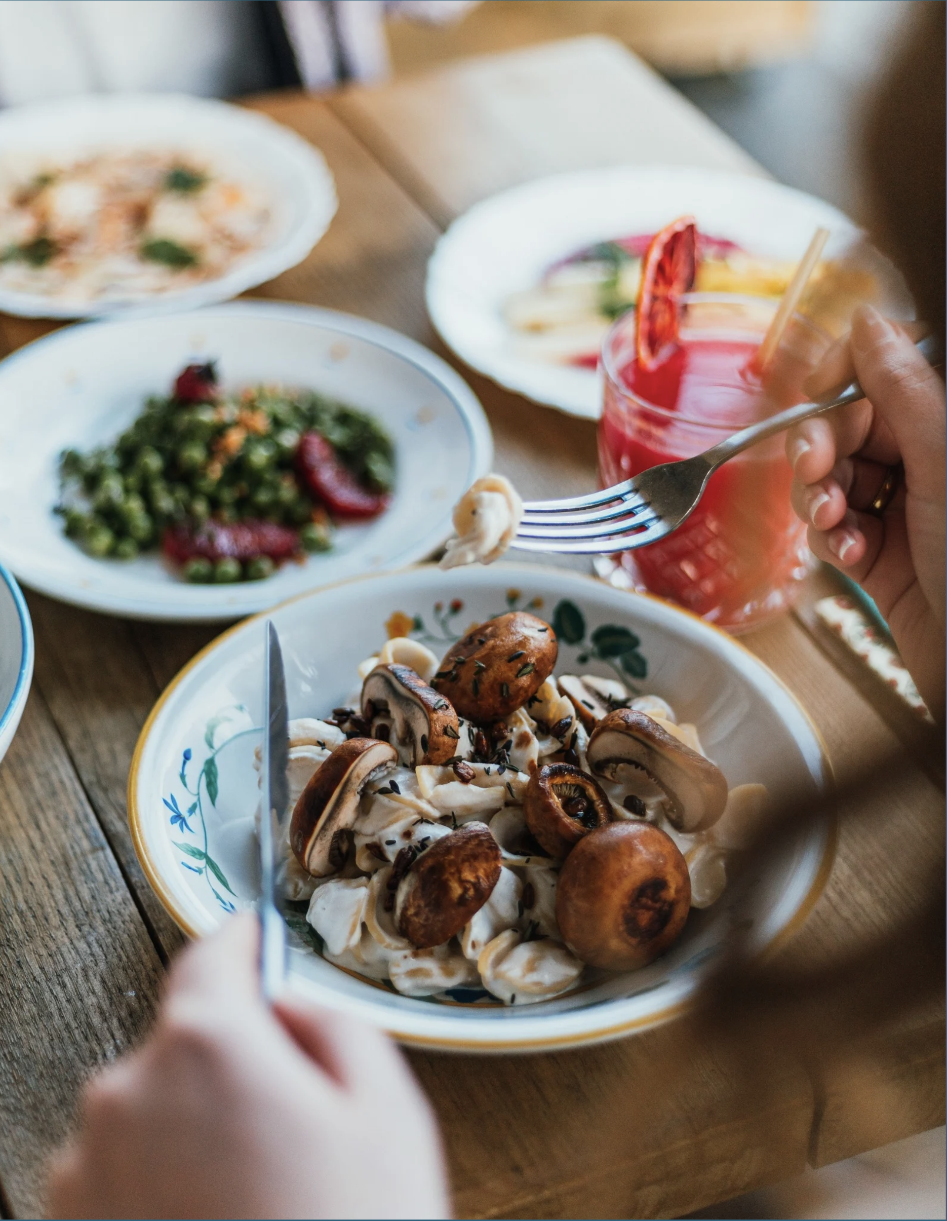 Gros plan sur un bol de champignons sautés avec des herbes, avec une fourchette dans une main, en arrière-plan d'autres plats, y compris une salade et une boisson rose avec une tranche de fruit.