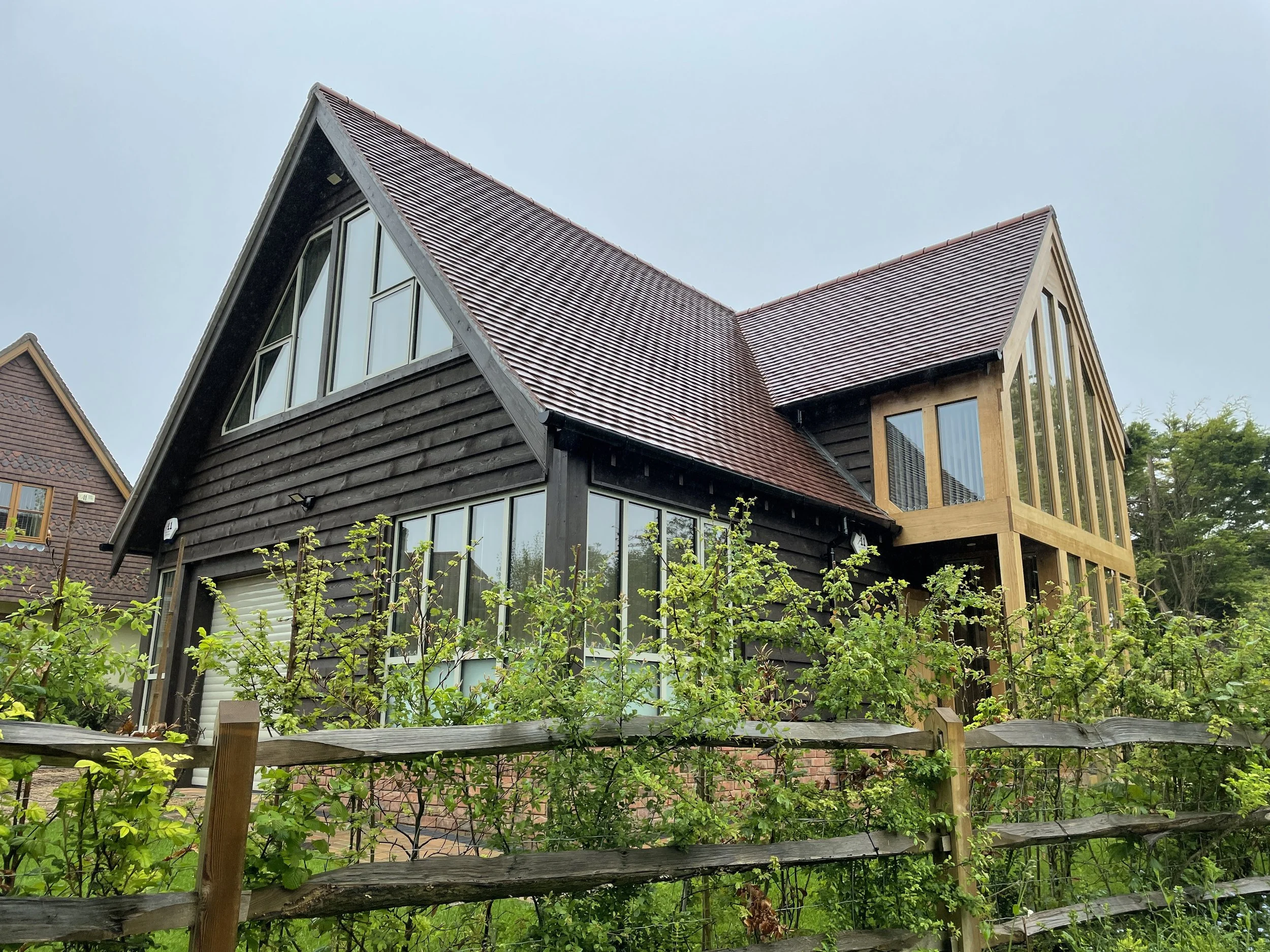 A modern house with dark wood siding and large windows, surrounded by greenery and a wooden fence.