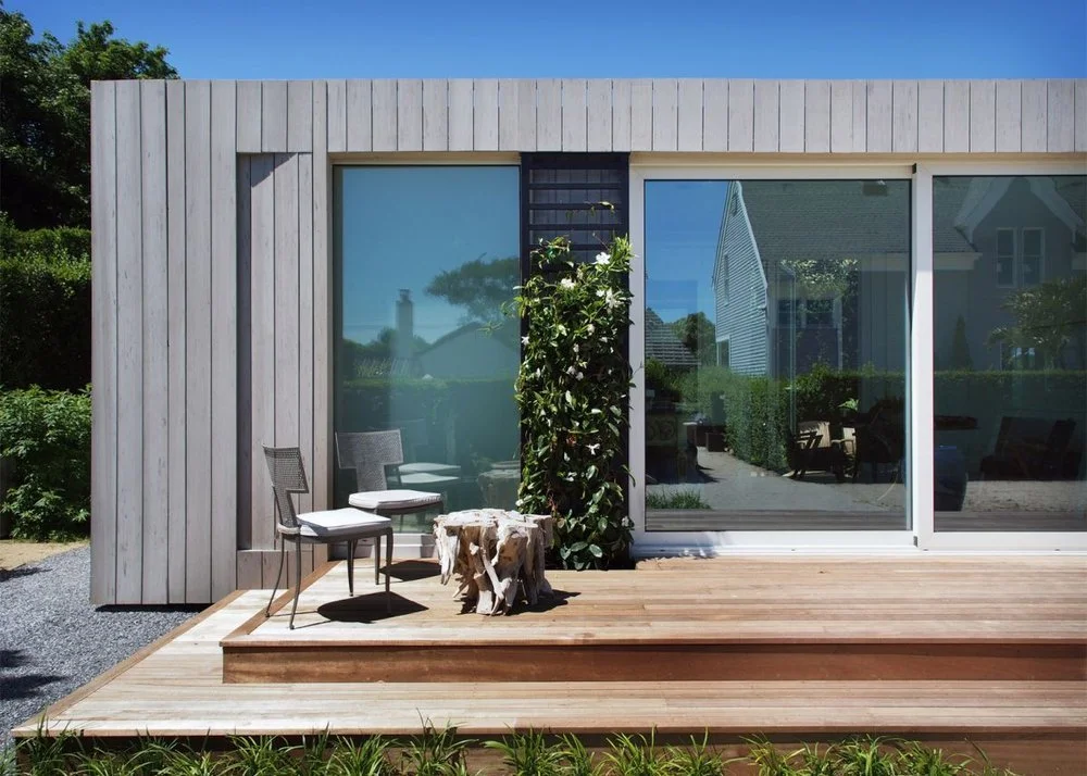 Modern house with a sliding glass door, a small wooden deck, two chairs, a tree stump, and green plants in the yard under a clear blue sky.