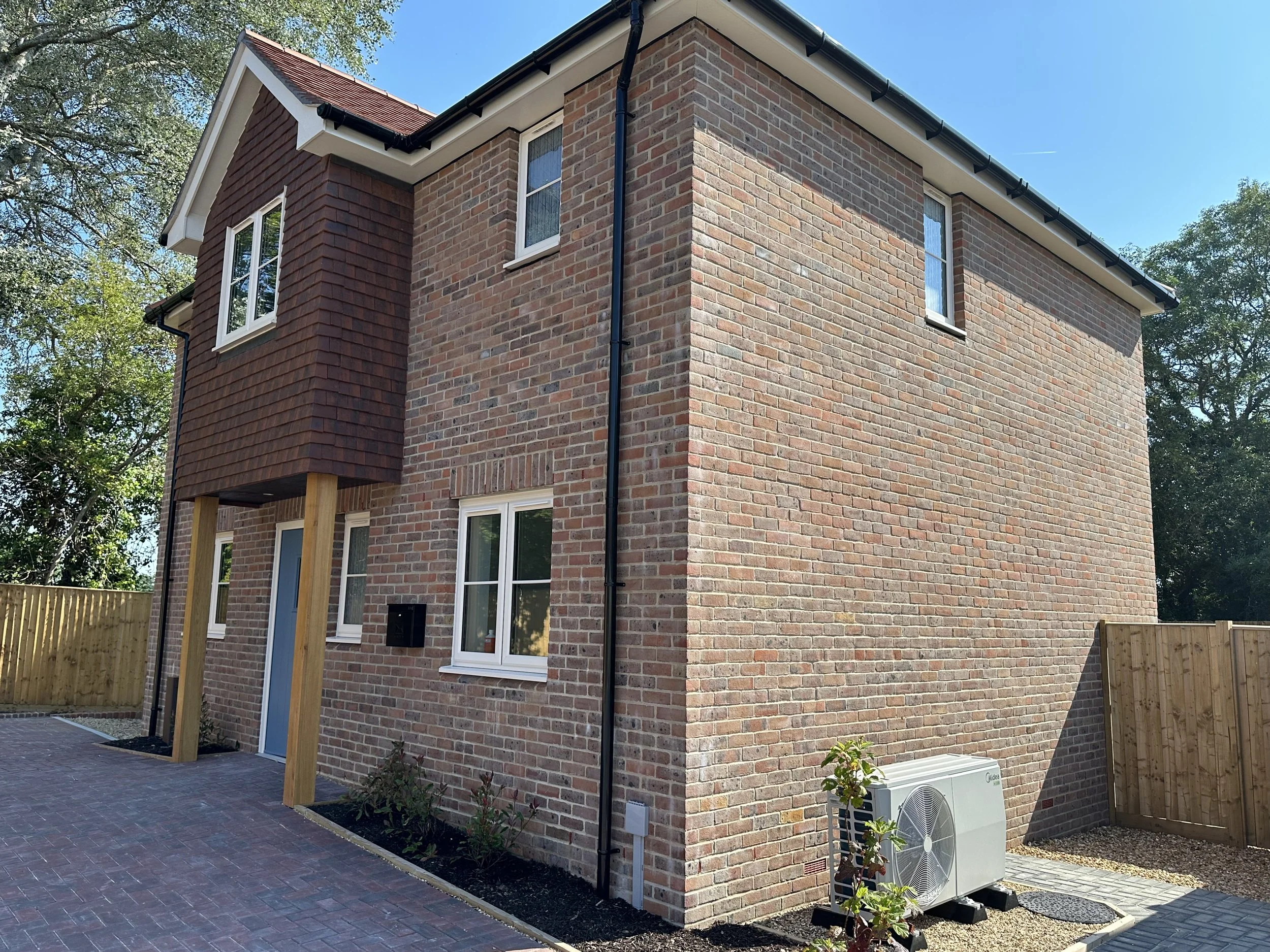 A modern two-story brick house with a brick and shingle exterior, white-framed windows, a blue front door, and a black gutter system. There is a small paved driveway, a wooden fence, and an outdoor air conditioning unit in the yard.