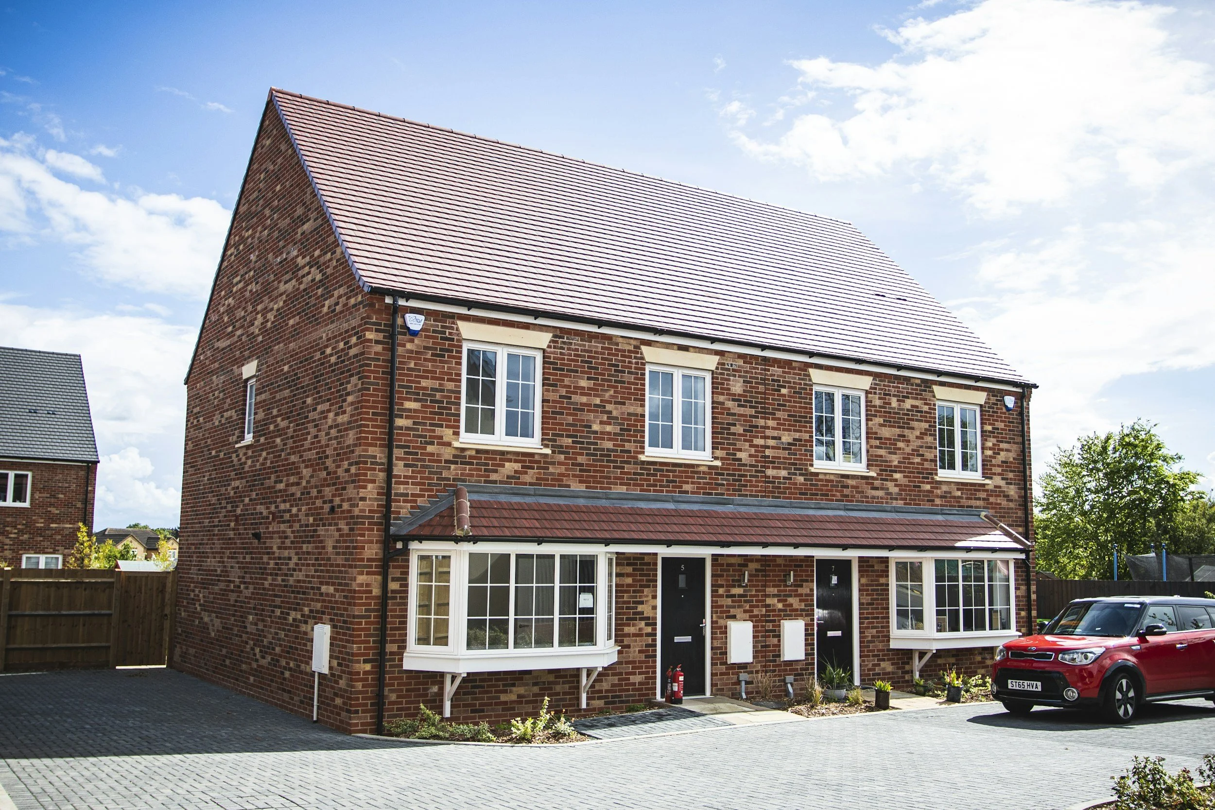 A modern brick residential building with a steep roof and white-framed windows, beside a paved driveway with a red car parked in front.