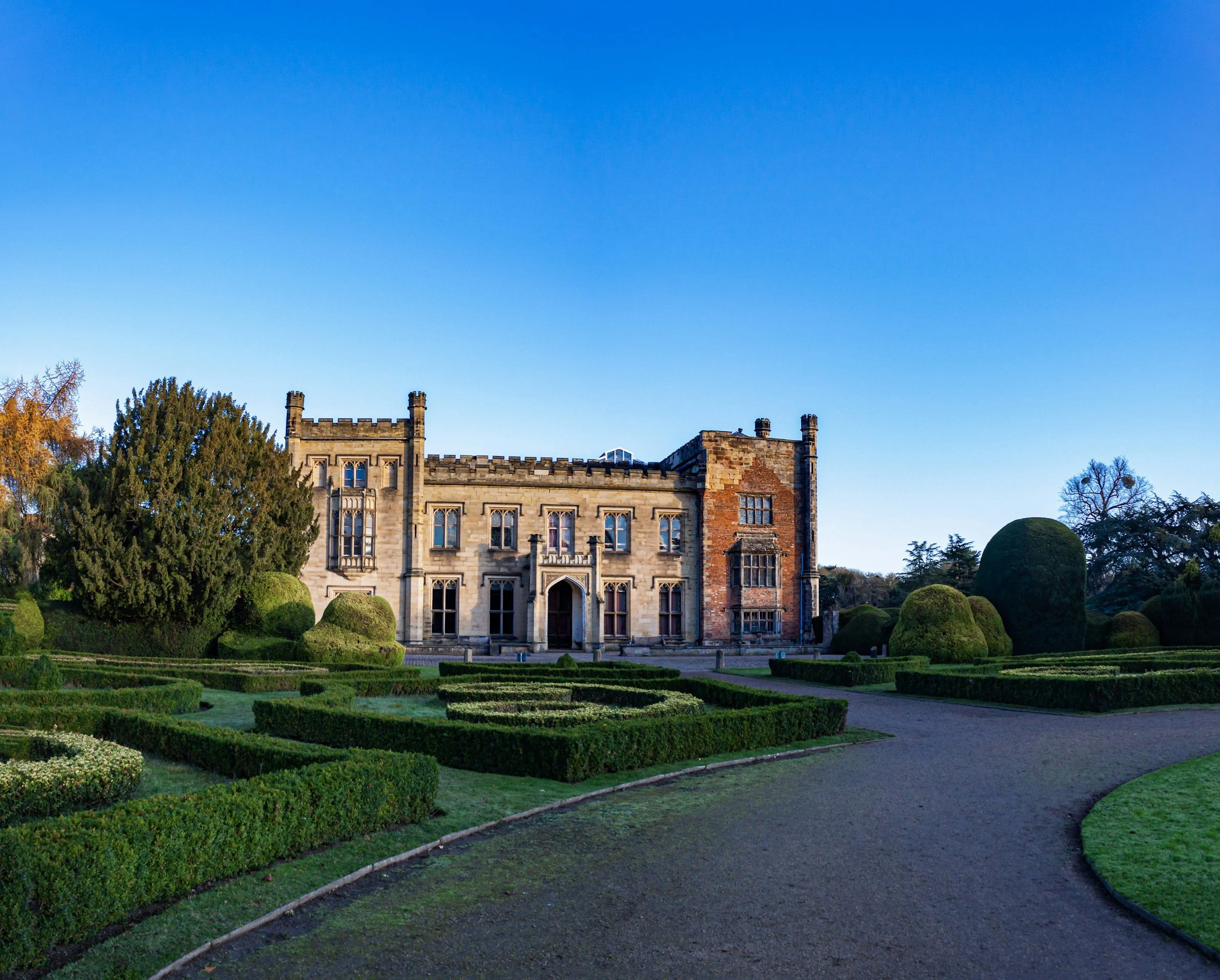 A large historic castle surrounded by neatly trimmed gardens and pathways under a clear blue sky.