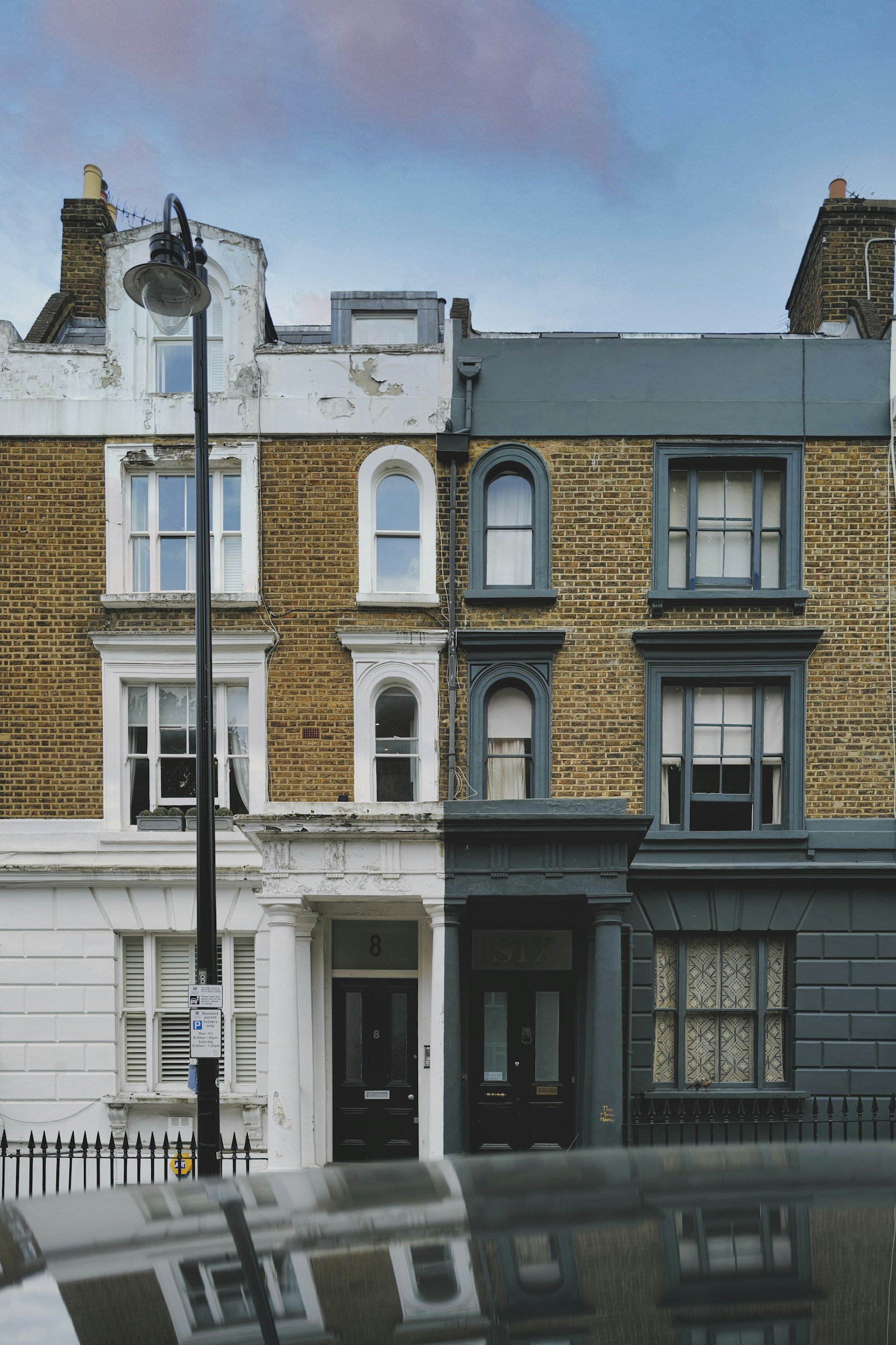 Multi-story brick townhouse with various window styles, a black door with the number 8, a streetlamp in front, and a reflection of the building in a car's hood at the bottom. The sky is partly cloudy.