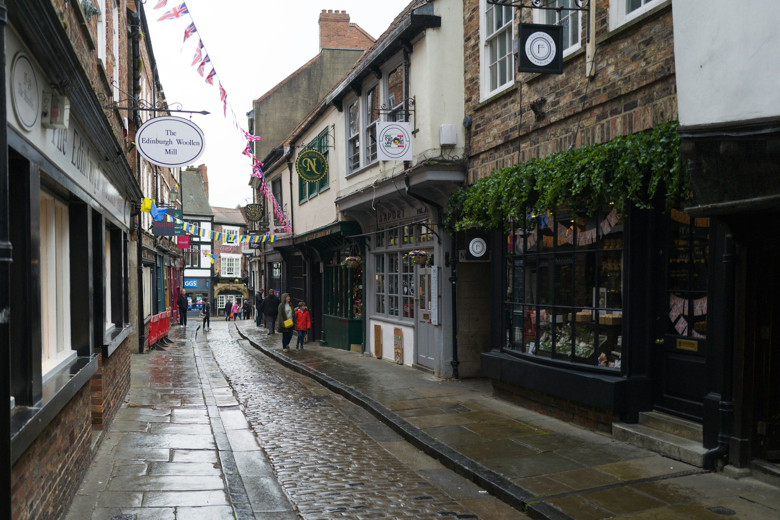 A narrow street in a historic town, wet from rain, with cobblestone and pavement, lined with shops and cafes, hanging colorful flags, and people walking.