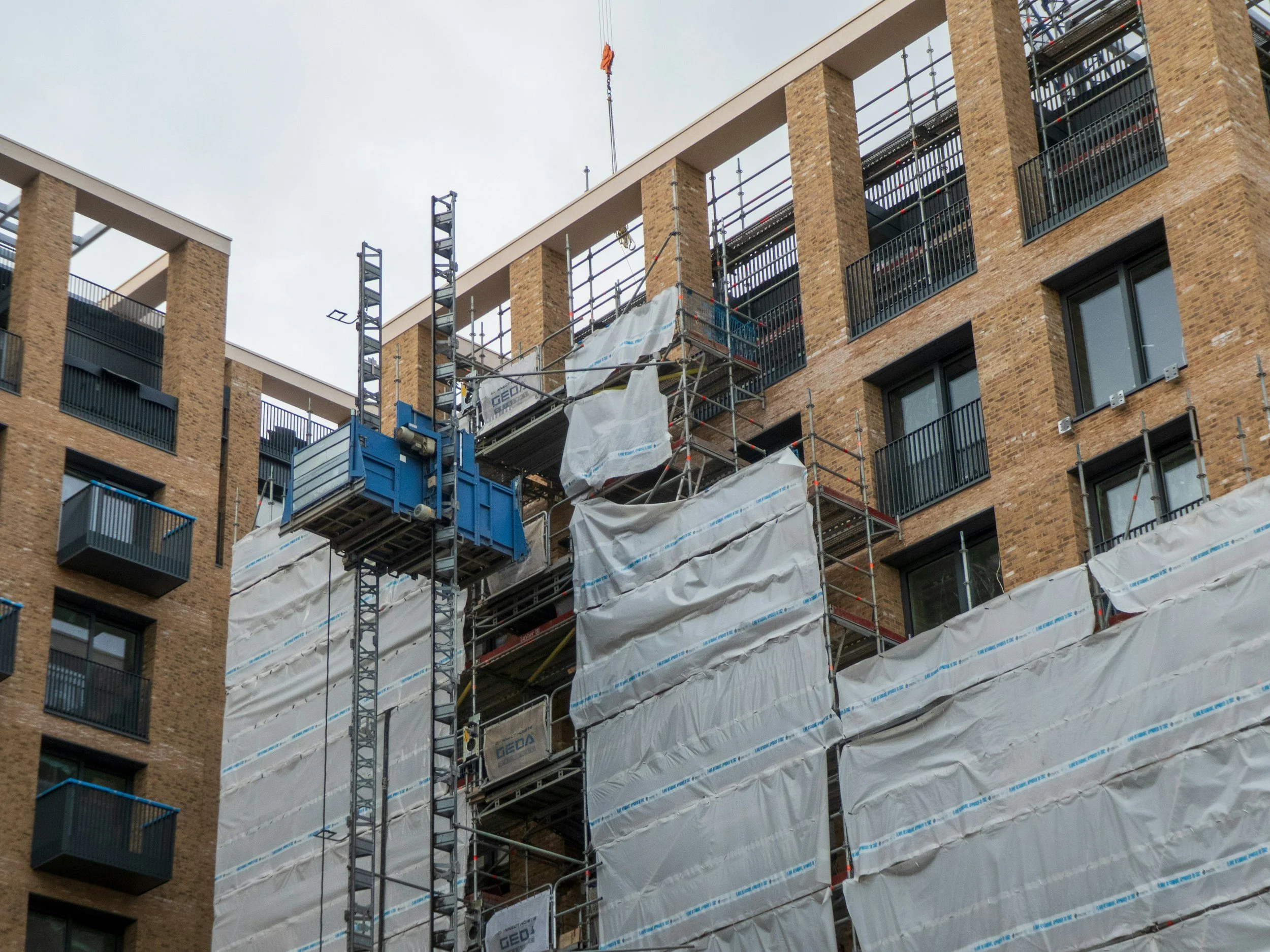 Construction site on a multi-story brick apartment building, with scaffolding and protective white sheeting covering part of the exterior walls.