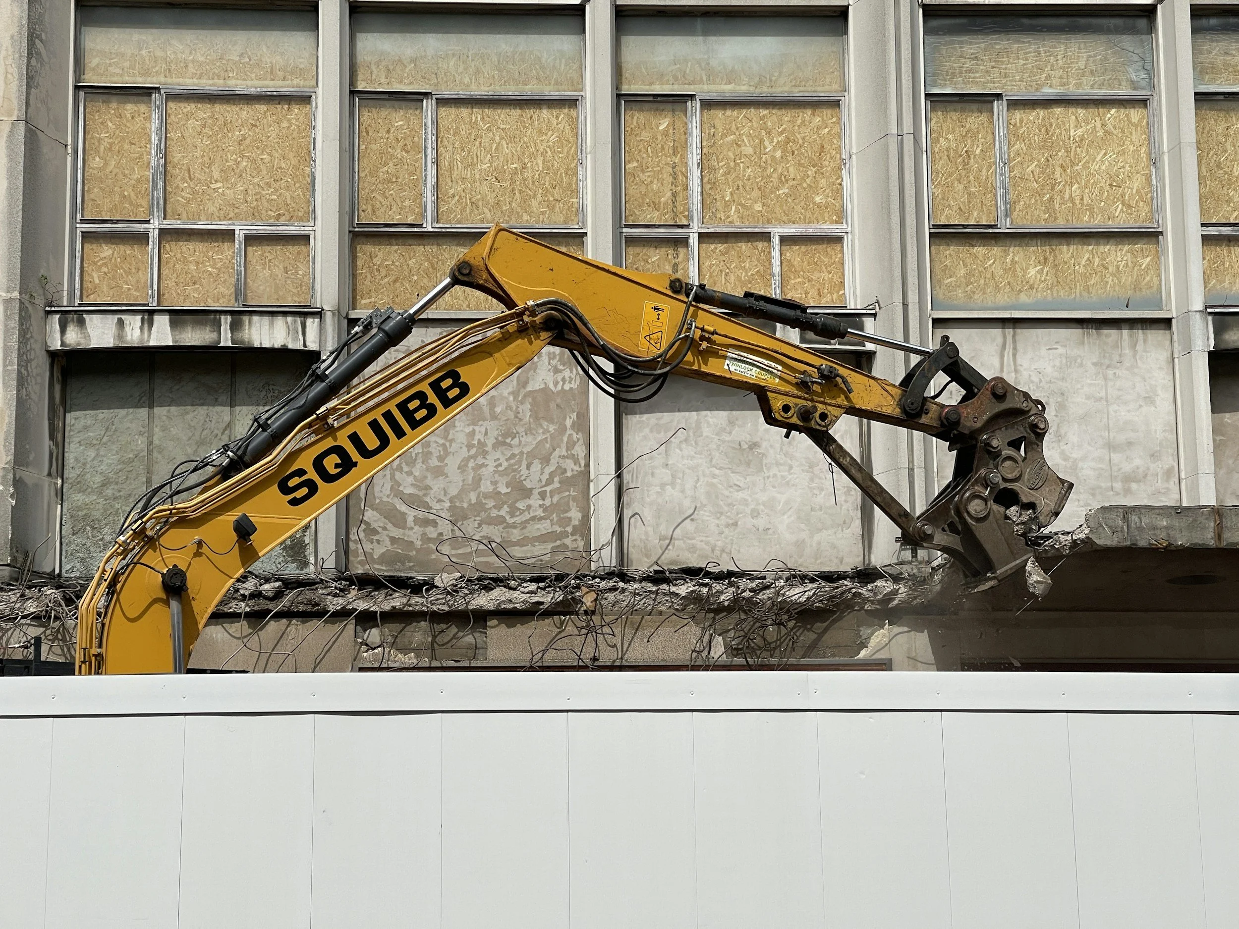 A yellow excavator demolishing an old building with boarded-up windows.