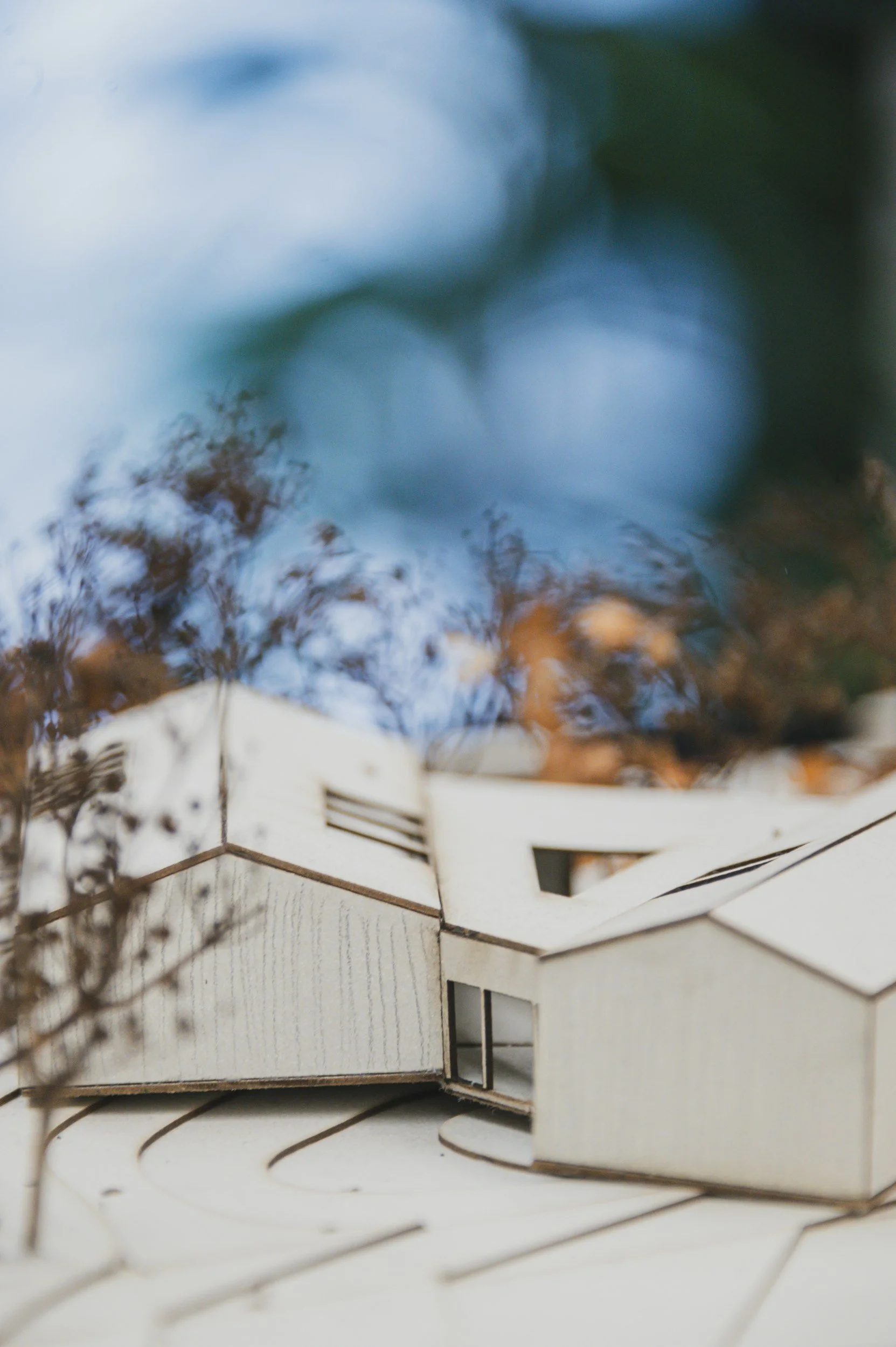 Close-up view of an architectural model made of light-colored wood or cardboard, with a modern design featuring angular shapes and large windows, situated outdoors with blurred trees and sky in the background.