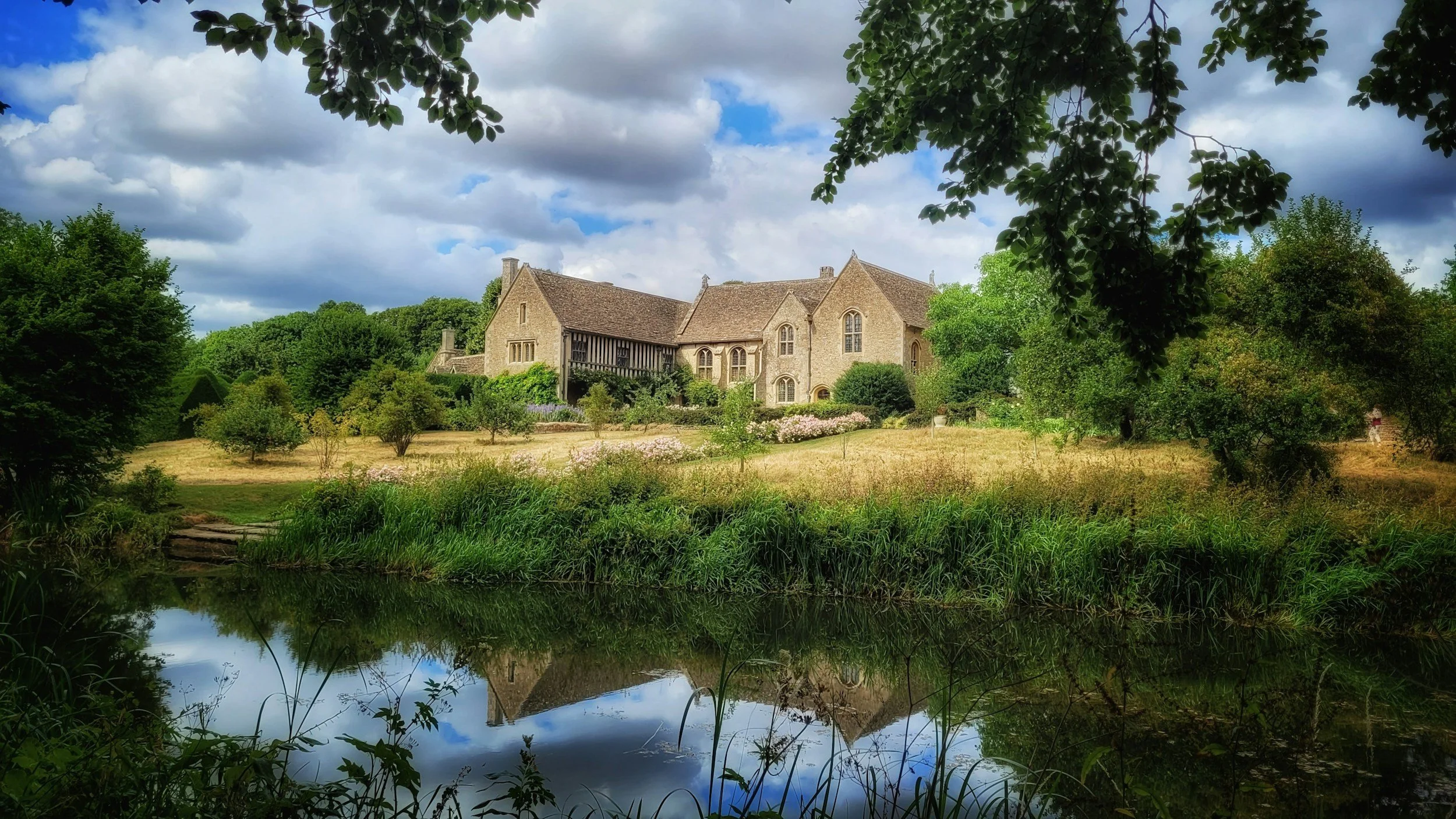A large stone house with a sloped roof, surrounded by trees and a garden, viewed from across a pond that reflects the scene. The sky is partly cloudy with patches of blue.