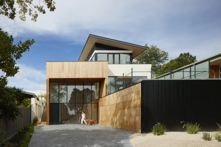 Modern multi-story house with wooden and black exterior finishes, large glass windows, and a driveway with a dog and a woman walking on it.