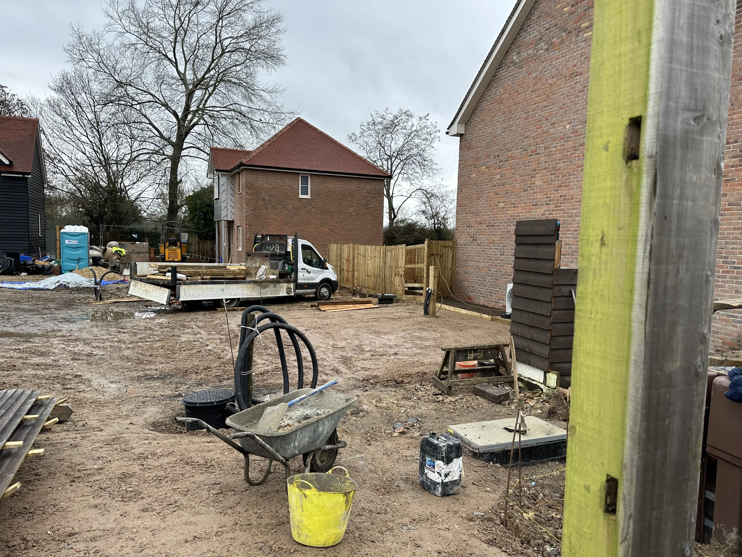 Construction site with a small truck, wheelbarrow, building materials, and workers, surrounded by partially built structures and residential houses.