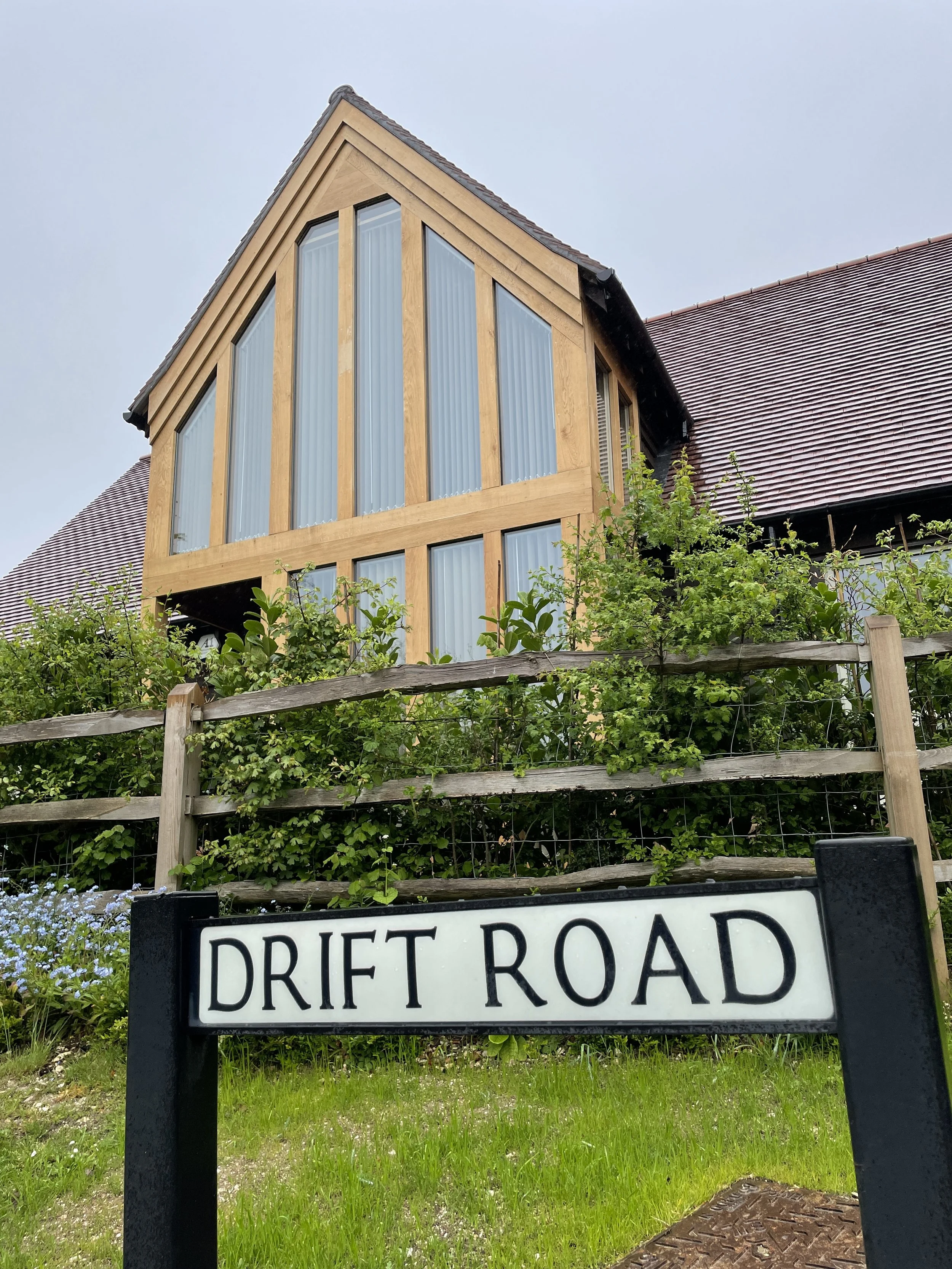 A house with large windows and a wooden exterior, a dark roof, surrounded by greenery, with a street sign that reads 'Drift Road' in the foreground.