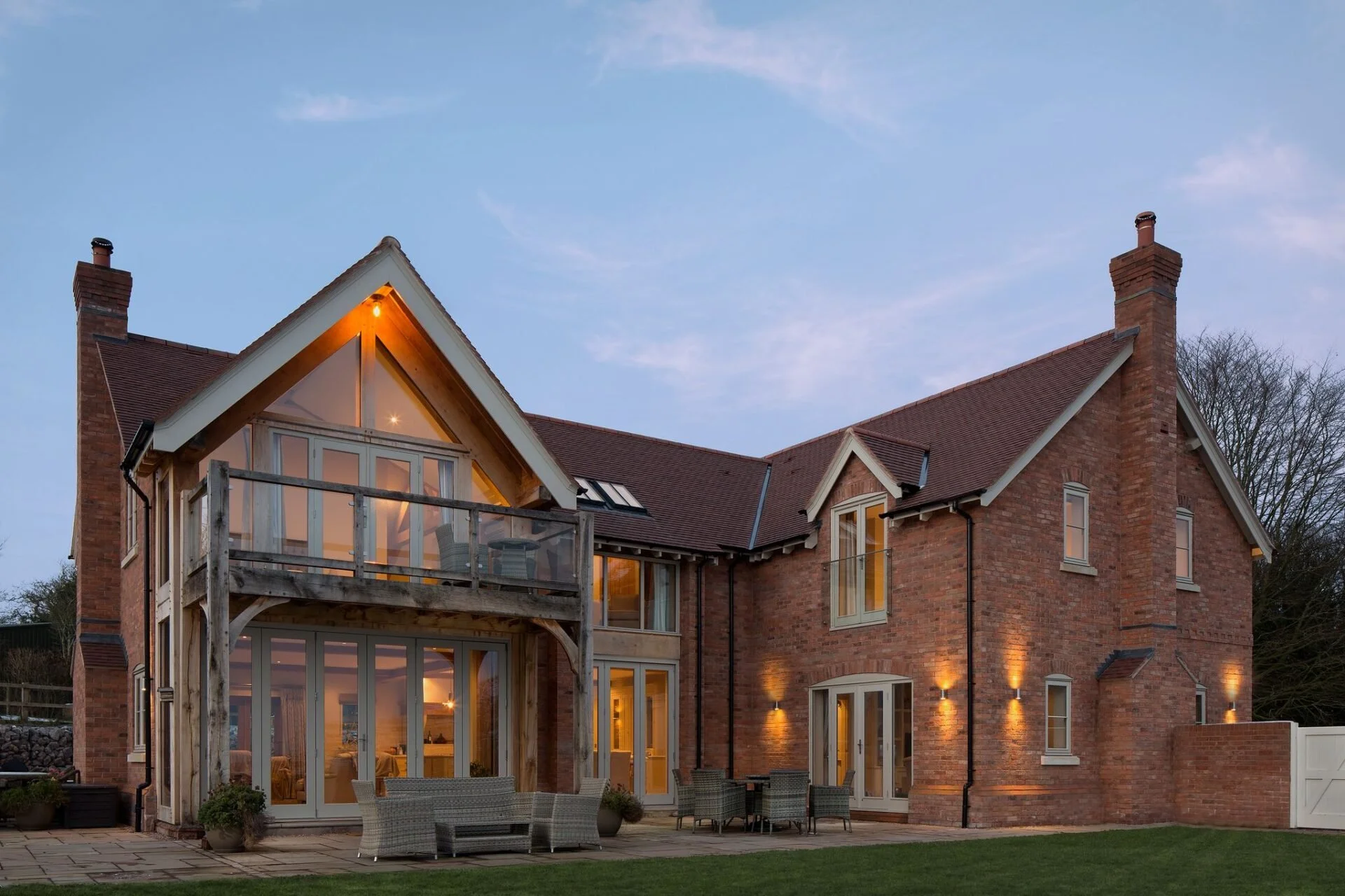 A brick house with a steeply pitched roof, illuminated windows, outdoor patio furniture, and a balcony, during dusk.