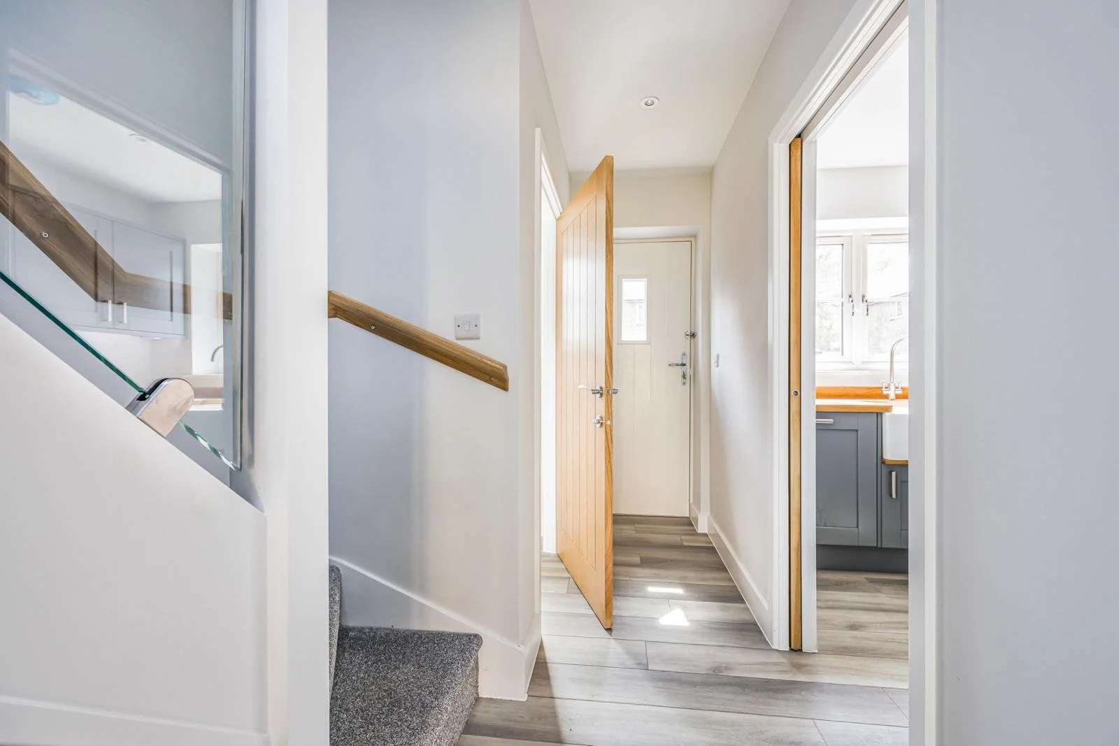 A hallway view showing a staircase leading to the upper floor, a wooden door slightly open, and a kitchen area with a window, bathroom sink, and gray cabinets visible through an open doorway.