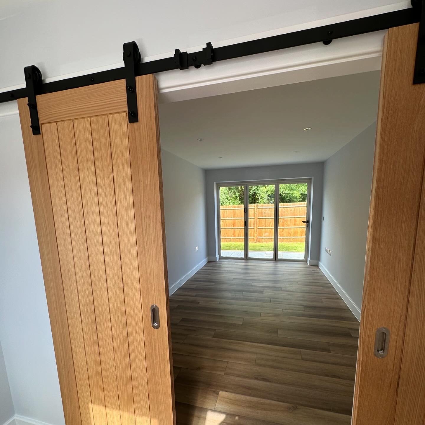 View of an empty room with glass sliding doors opening to a backyard with a wooden fence, hardwood floor, white walls, and recessed lighting, seen through a partially open wooden sliding door.
