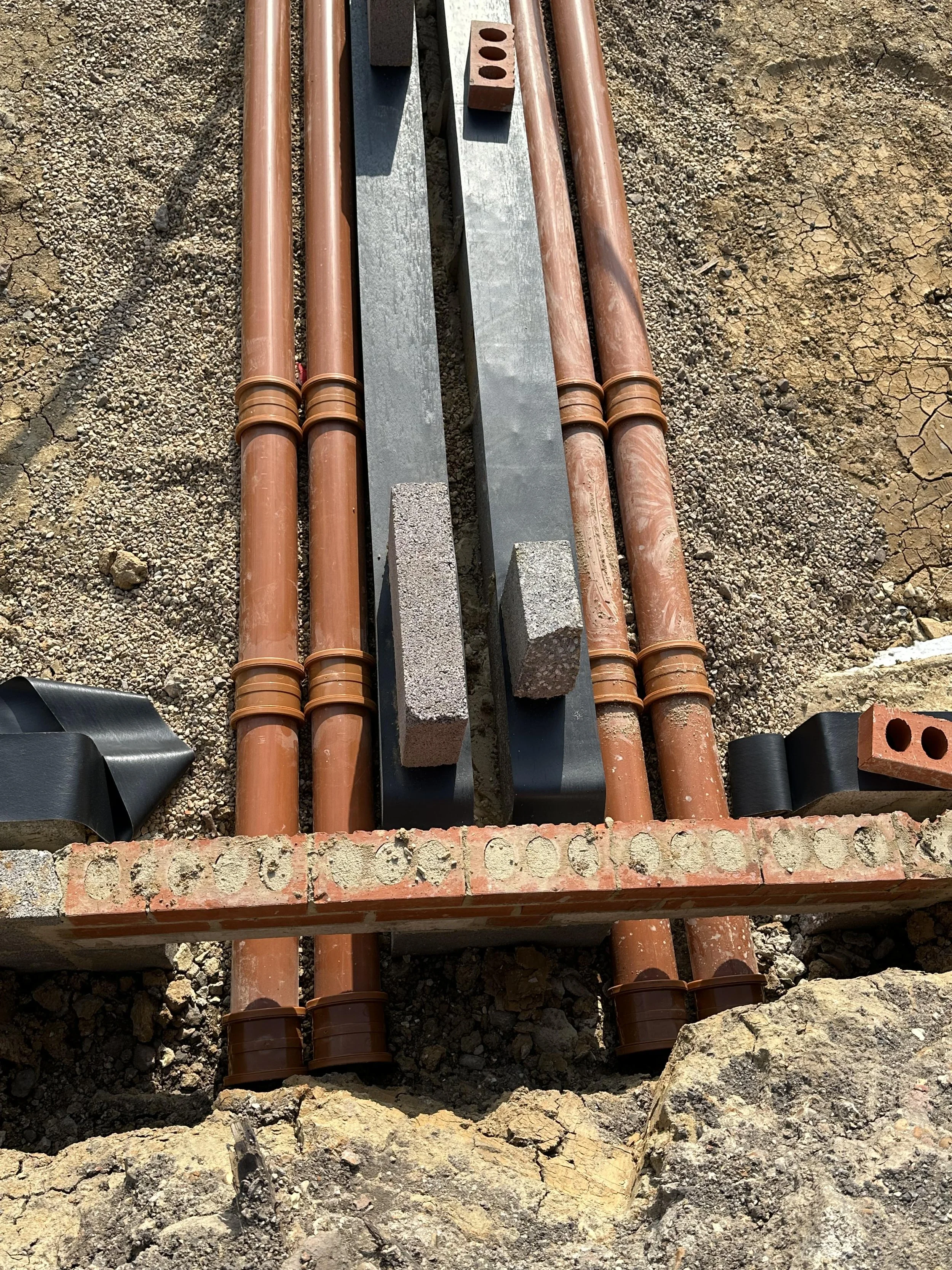 Construction site with exposed orange PVC pipes, bricks, metal beams, and gravel ground.