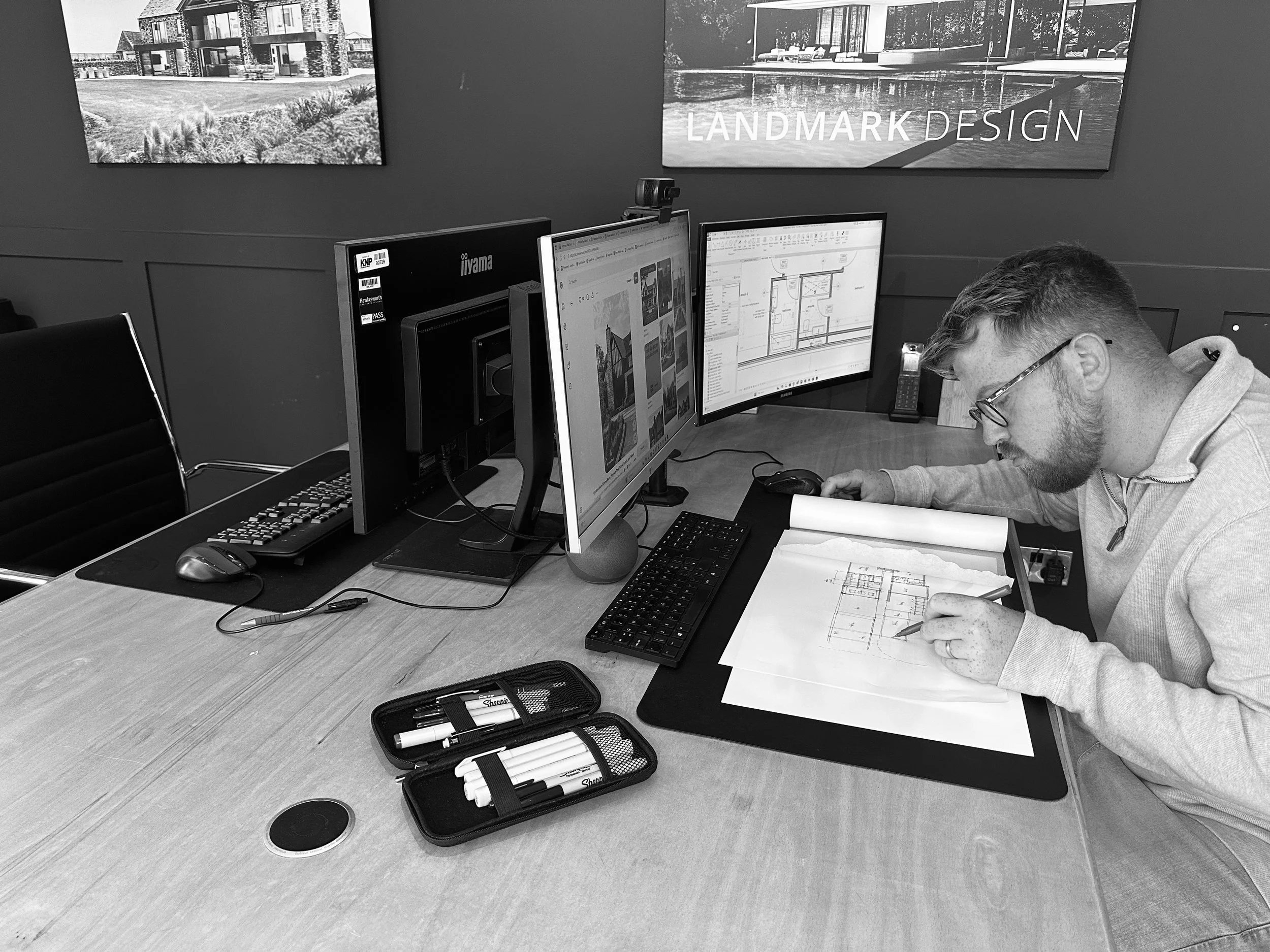 A man wearing glasses is working on architectural blueprints at a desk with three computer monitors. The workspace has a poster on the wall that reads "LANDMARK DESIGN." The desk also has a keyboard, mouse, phone, and a pencil case.