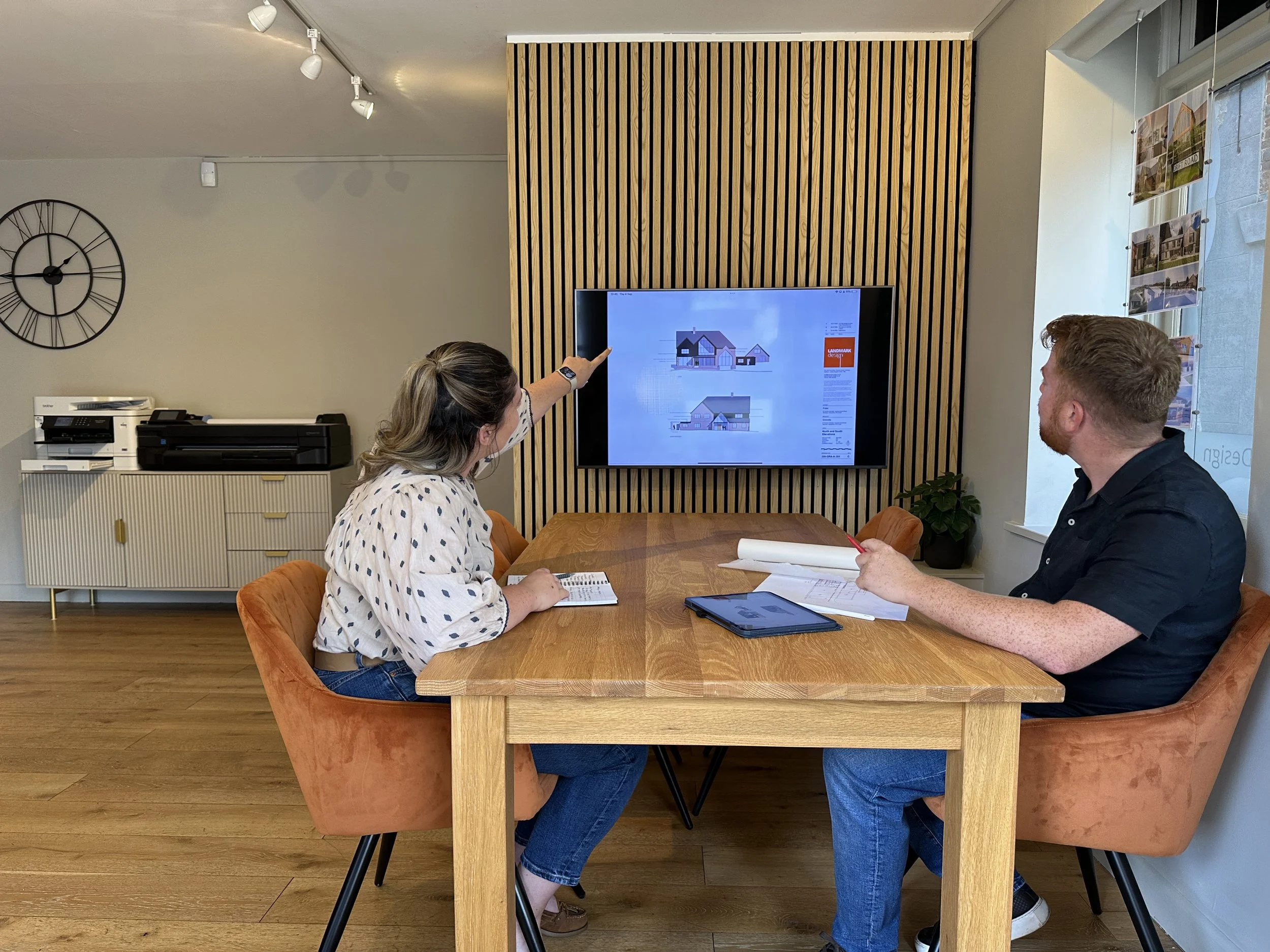 Two people sitting at a wooden table in an office, one woman and one man, with the woman pointing at a screen displaying architectural plans, while the man looks on and takes notes.
