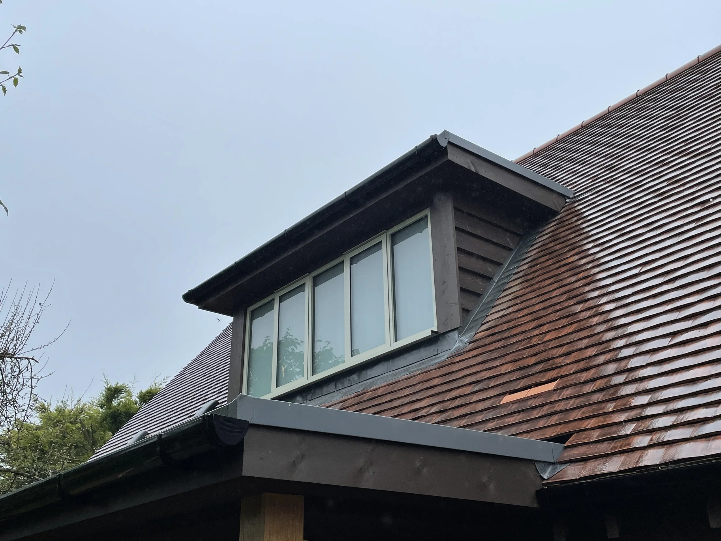 Close-up of a house roof with a dormer window, brown tiles, and black trim, with a cloudy sky in the background.