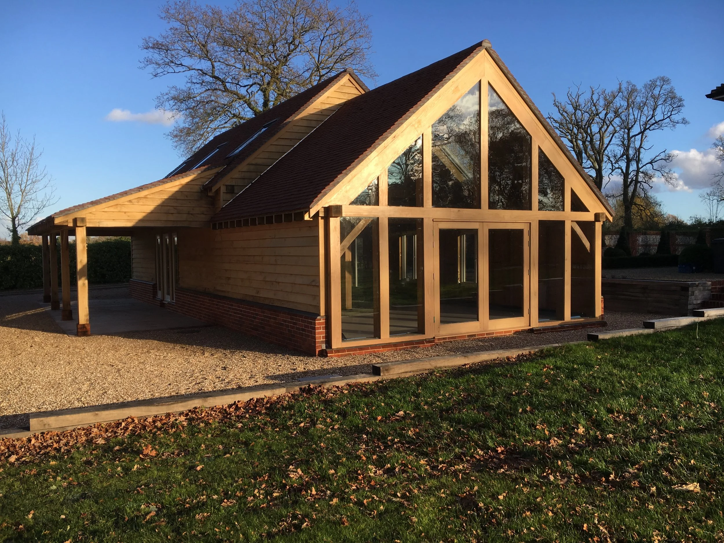 A building under construction with wooden framing and large glass windows, set in a grassy area with trees and a blue sky in the background.