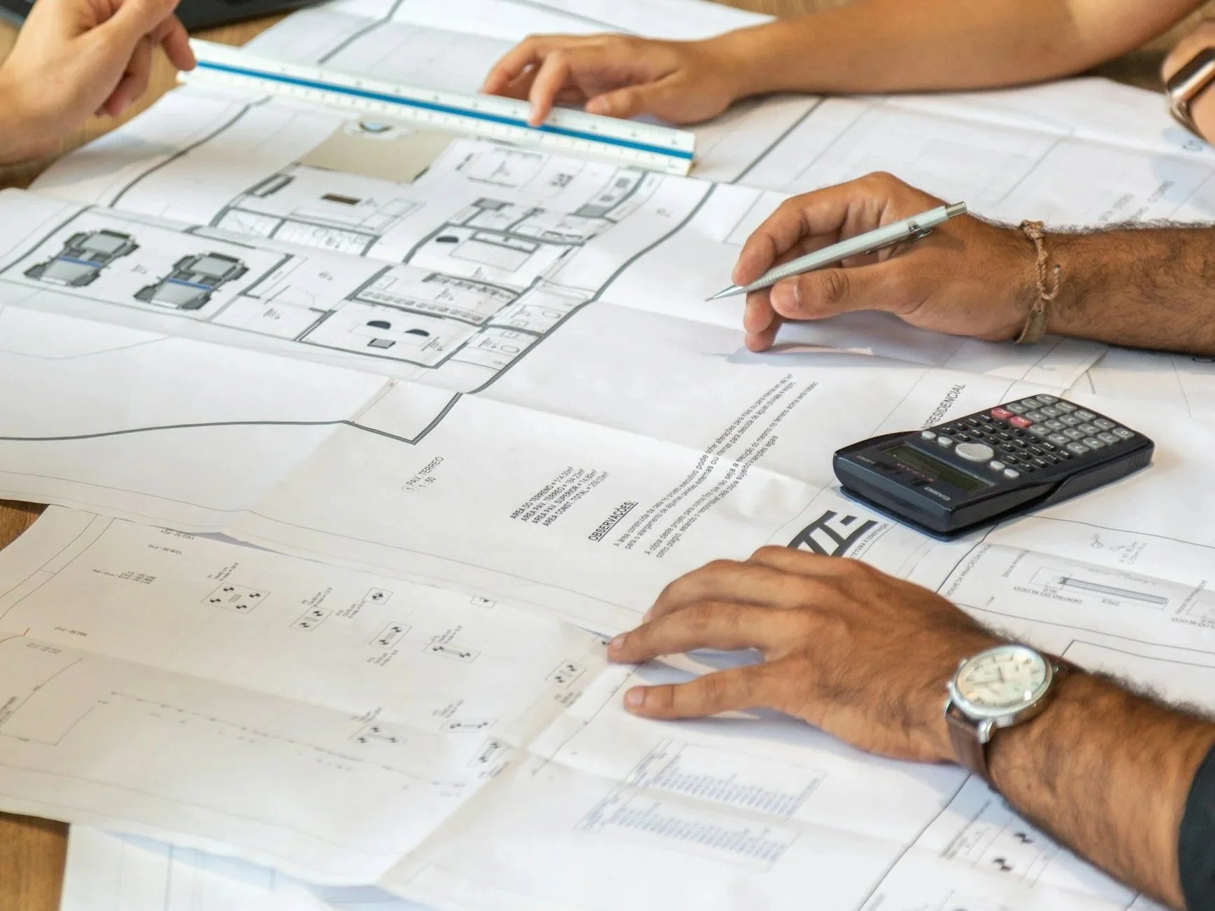Architectural plans and blueprints on a table with hands pointing and holding a pen, a calculator, and a scale.