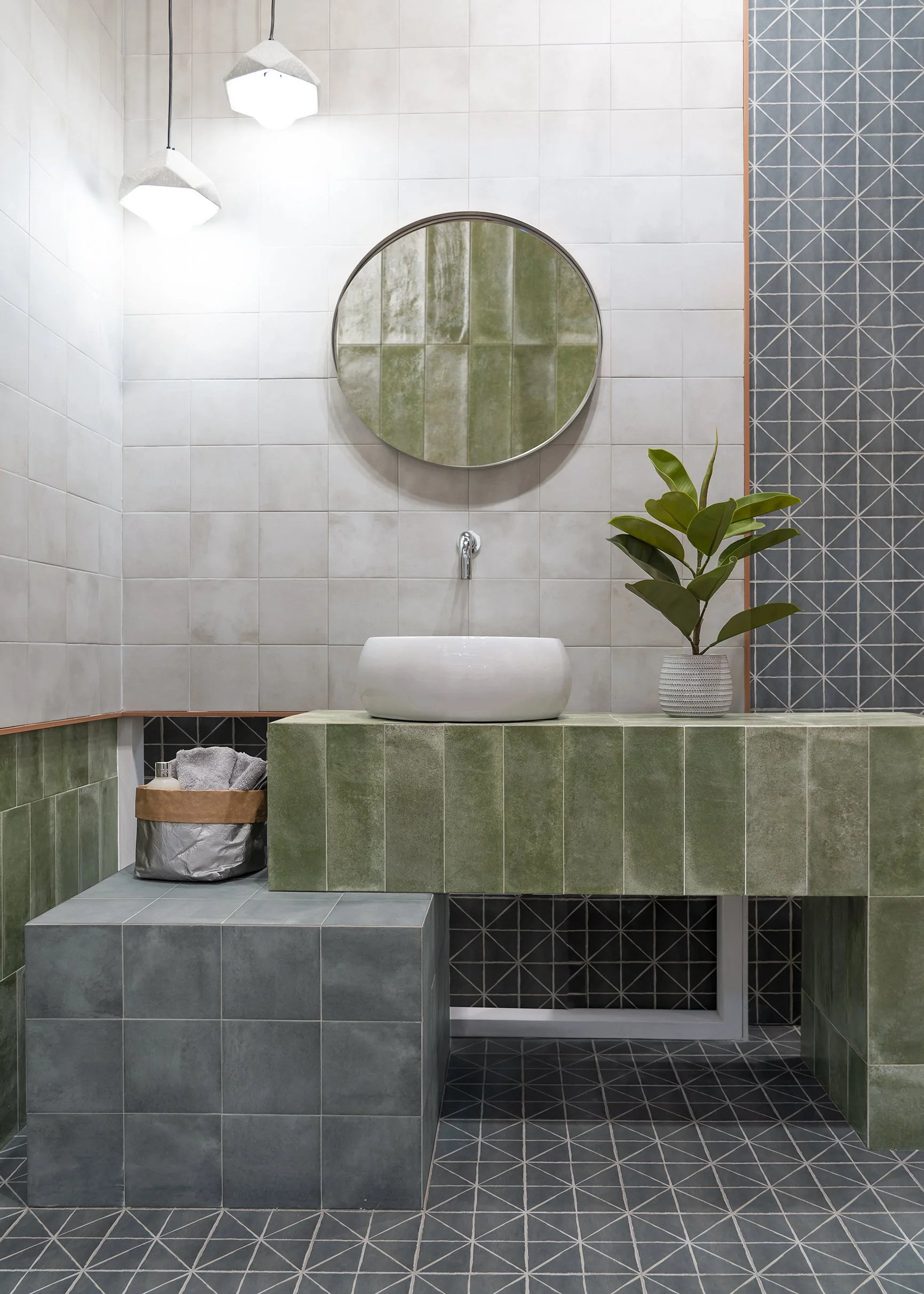 Modern bathroom with green and dark tile accents, a circular mirror, a white vessel sink, a potted plant, and a basket with towels.