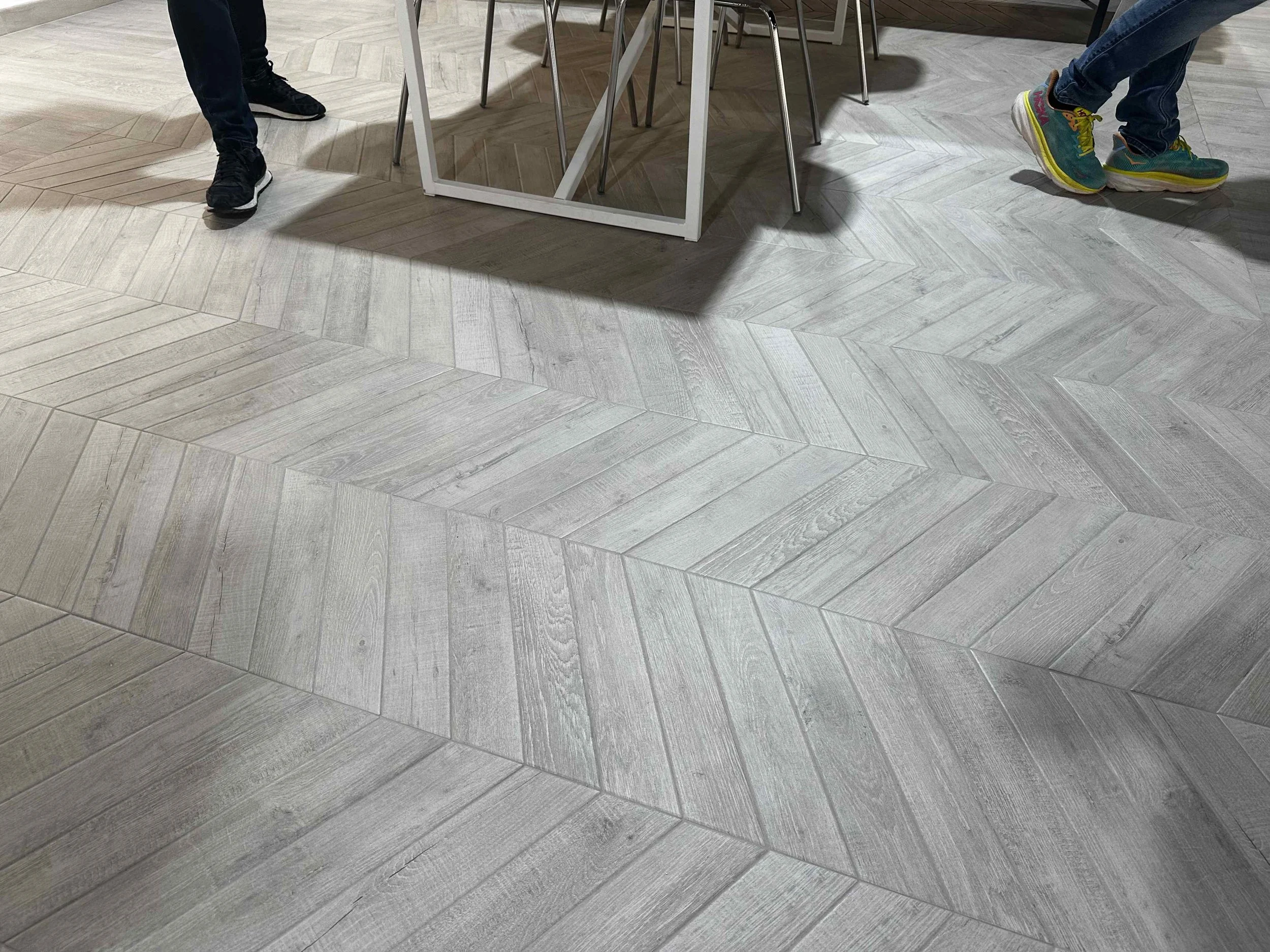 Close-up view of a modern, light-colored wooden flooring with a herringbone pattern, with two pairs of legs visible in a room with a table and chairs.