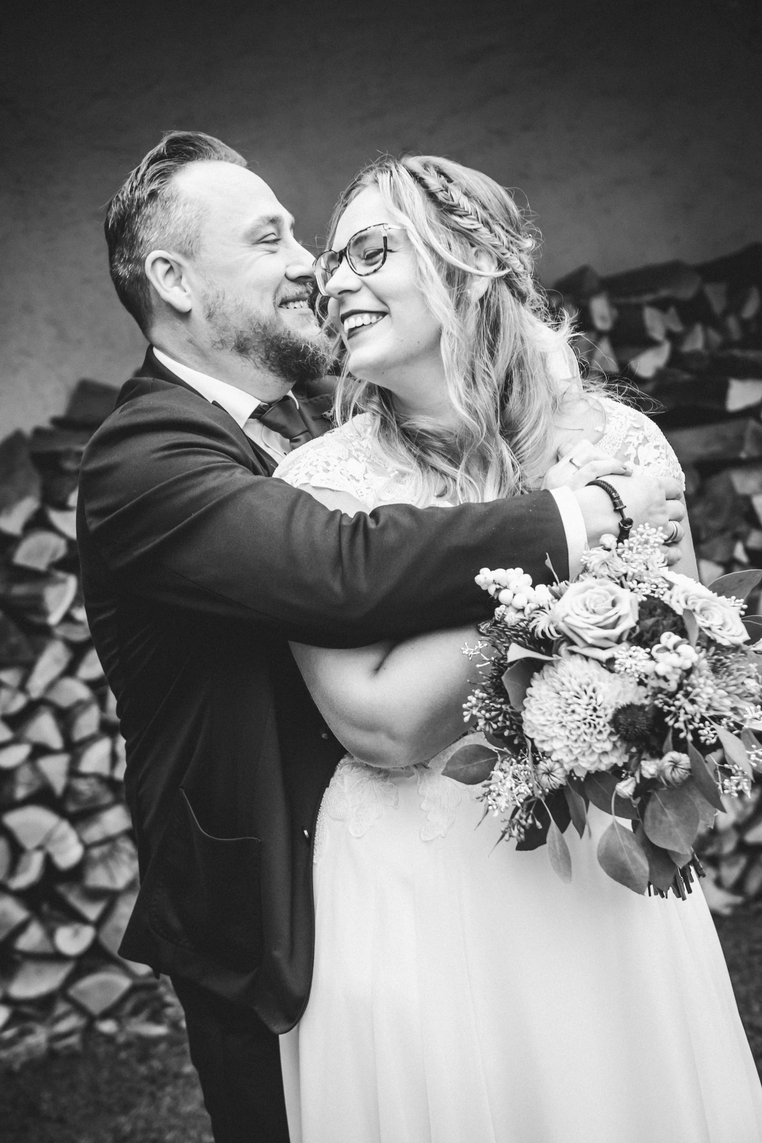 Ein glückliches Brautpaar bei einer Hochzeit, umarmend, im schwarzen und weißen Bild, mit einem Holzstapel im Hintergrund.
