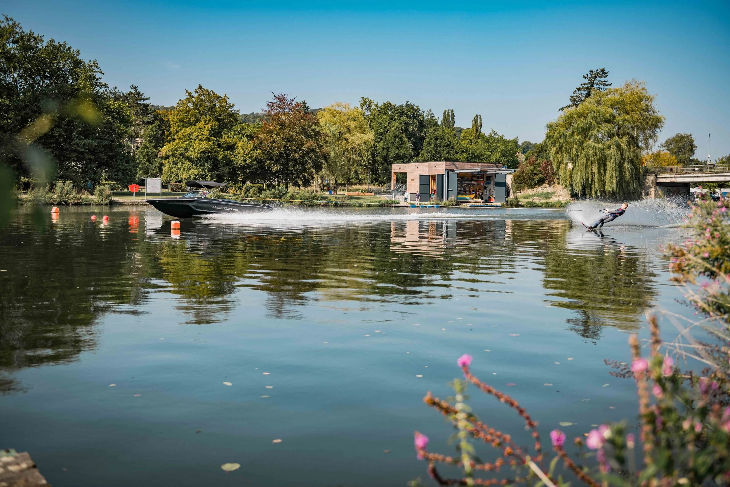 Ein Boot, das auf dem Wasser fährt, während eine Person Wasserski fährt. Im Hintergrund sind Bäume, eine kleine Hütte und eine Brücke sichtbar.
