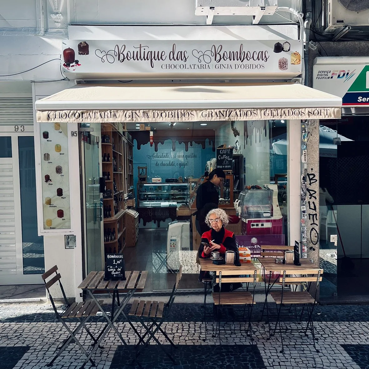 Ein kleines Café mit Sitzgelegenheiten vor dem Laden, einer älteren Frau, die an einem Tisch liest, und einem Mitarbeitenden im Inneren. Das Café verkauft Schokoladentorten, gefüllte und gelierte Süßigkeiten, sowie weiche Eiscremes, wie auf den Schil