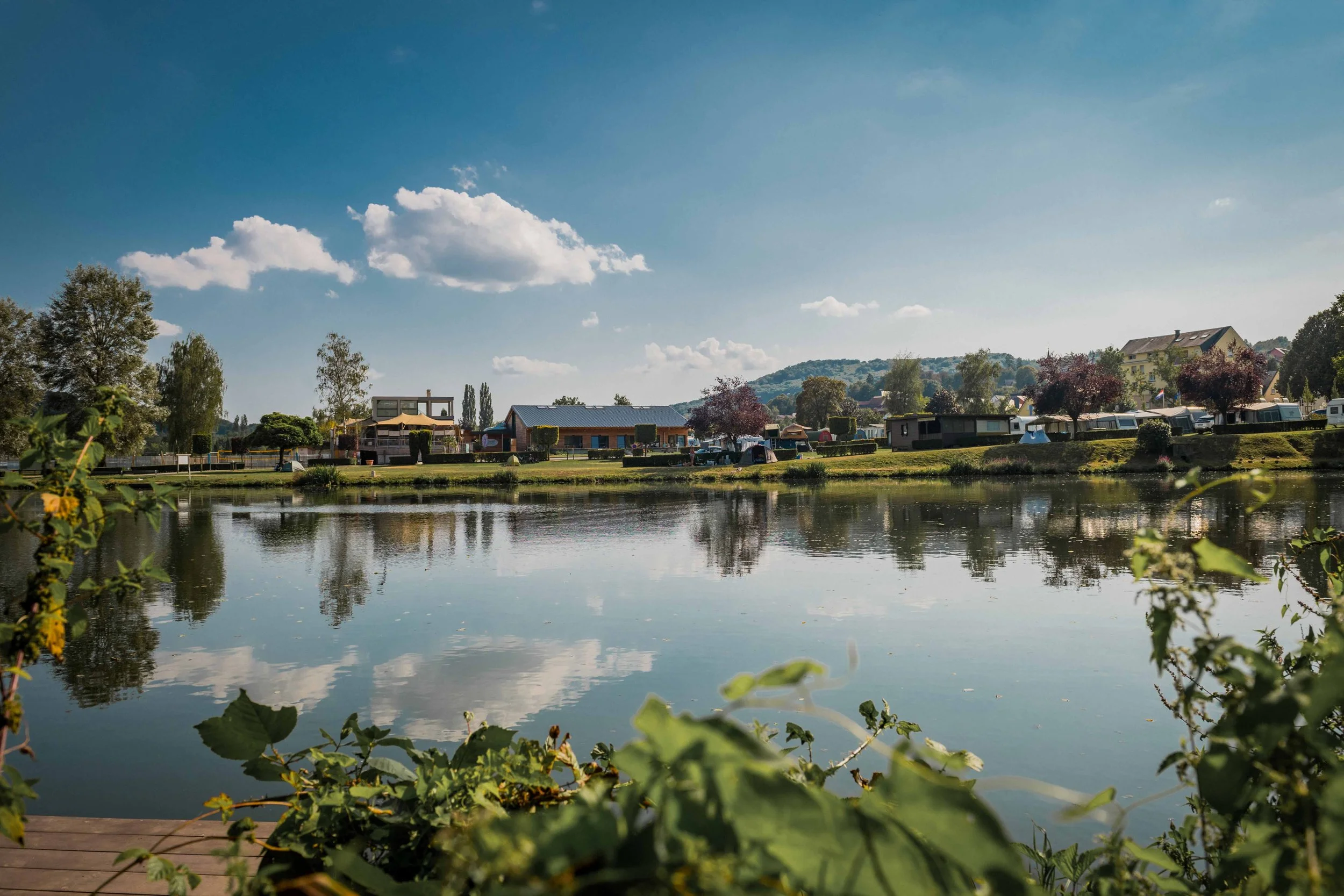 Blick auf einen ruhigen See mit Häusern und Bäumen im Hintergrund bei sonnigem Himmel, reflektiert im Wasser.