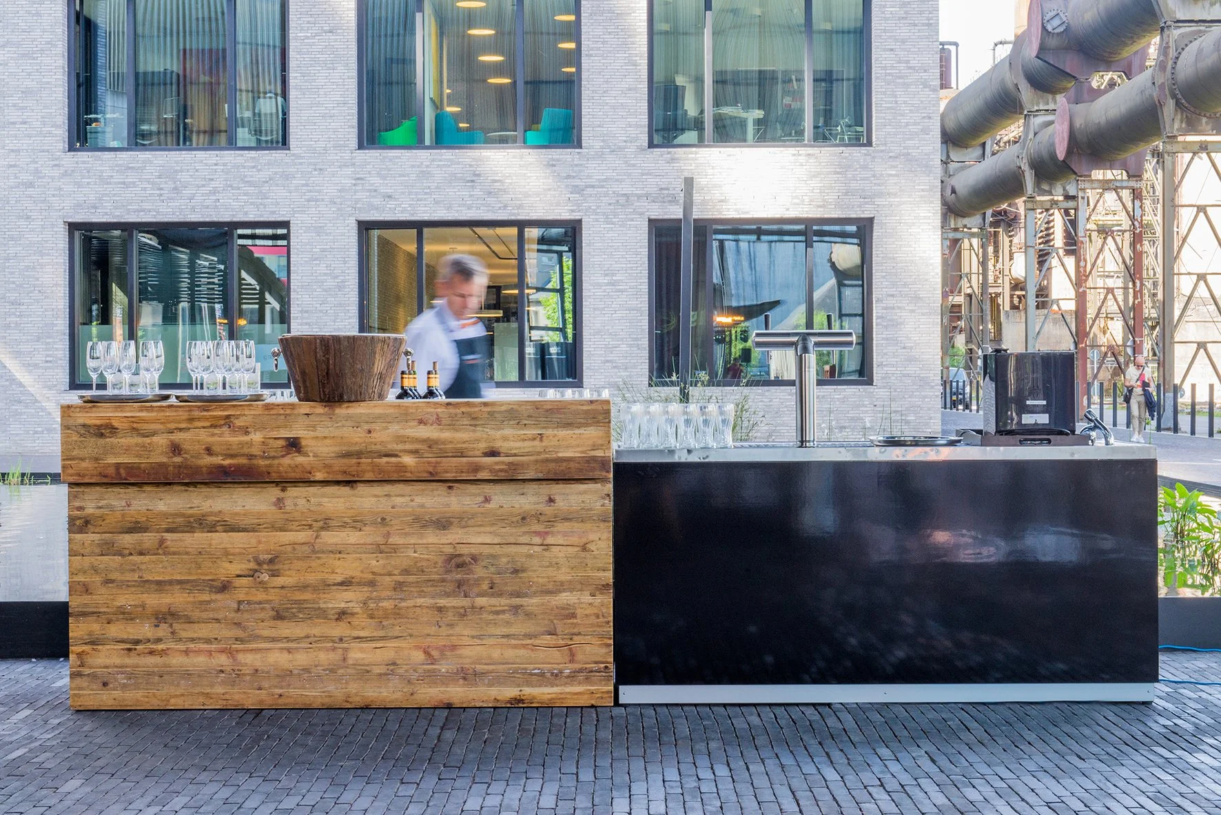 Ein Außenbar mit Holztresen, Glaswaren und einem Barkeeper vor einem modernen Gebäude mit großen Fenstern und sichtbaren Stadtinfrastruktur.