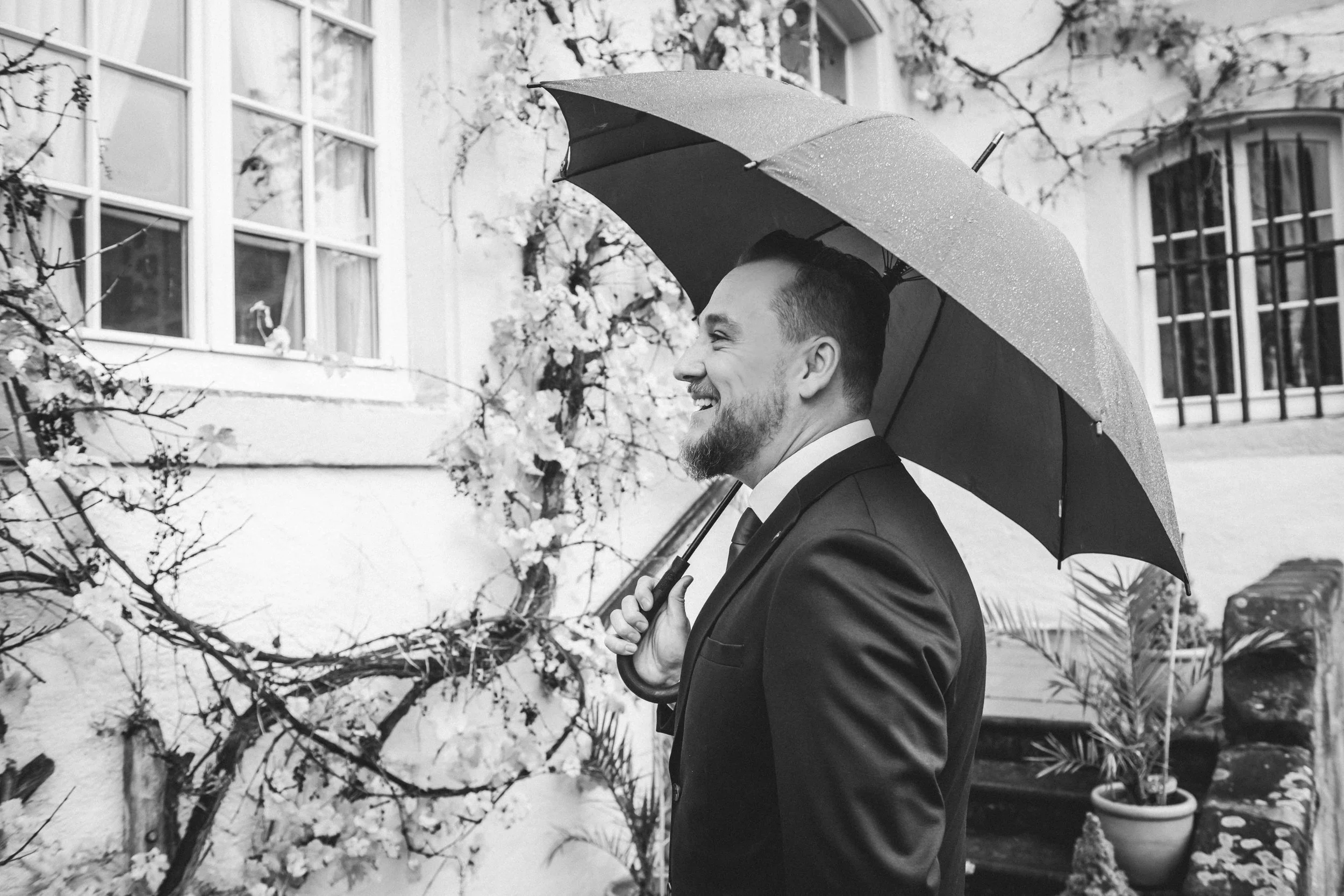 Ein lachender Mann in formeller Kleidung hält einen Regenschirm und steht im Freien vor einem Haus mit Blumen und Pflanzen.