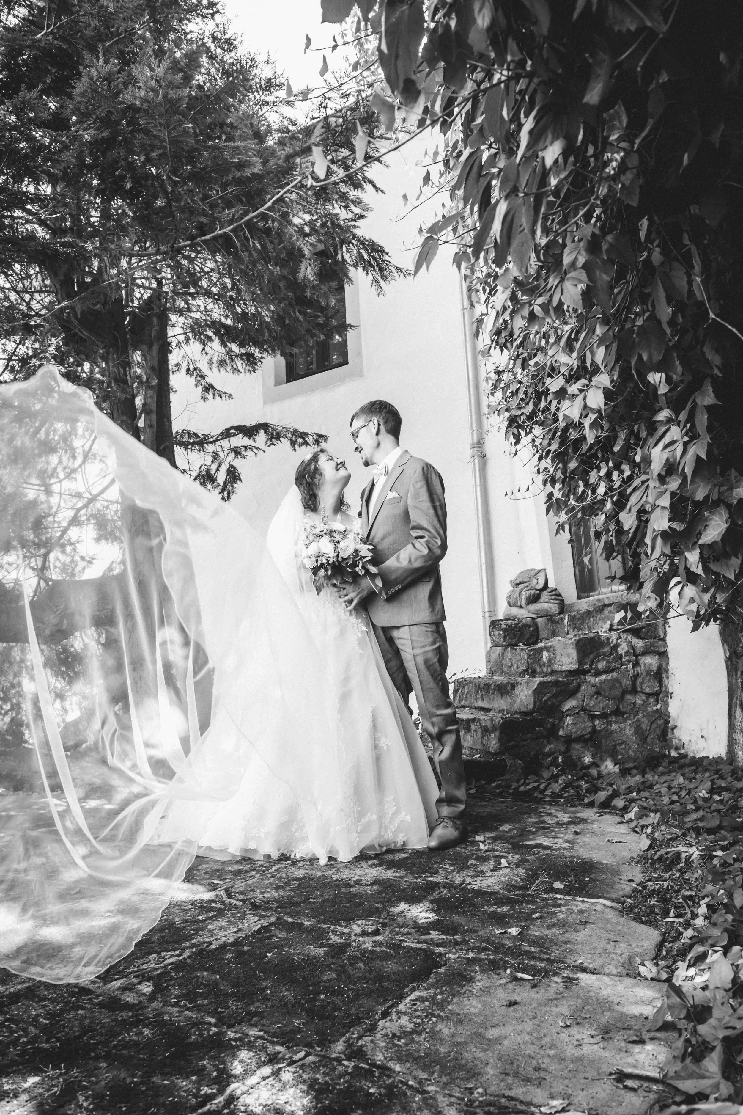 Ein Brautpaar steht im Freien vor einem weißen Haus, blickt sich liebevoll an und lächelt. Die Braut trägt ein Hochzeitskleid und hält einen Blumenstrauß, der Bräutigam trägt einen Anzug. Die Szene ist schwarz-weiß fotografiert, umgeben von Bäumen un
