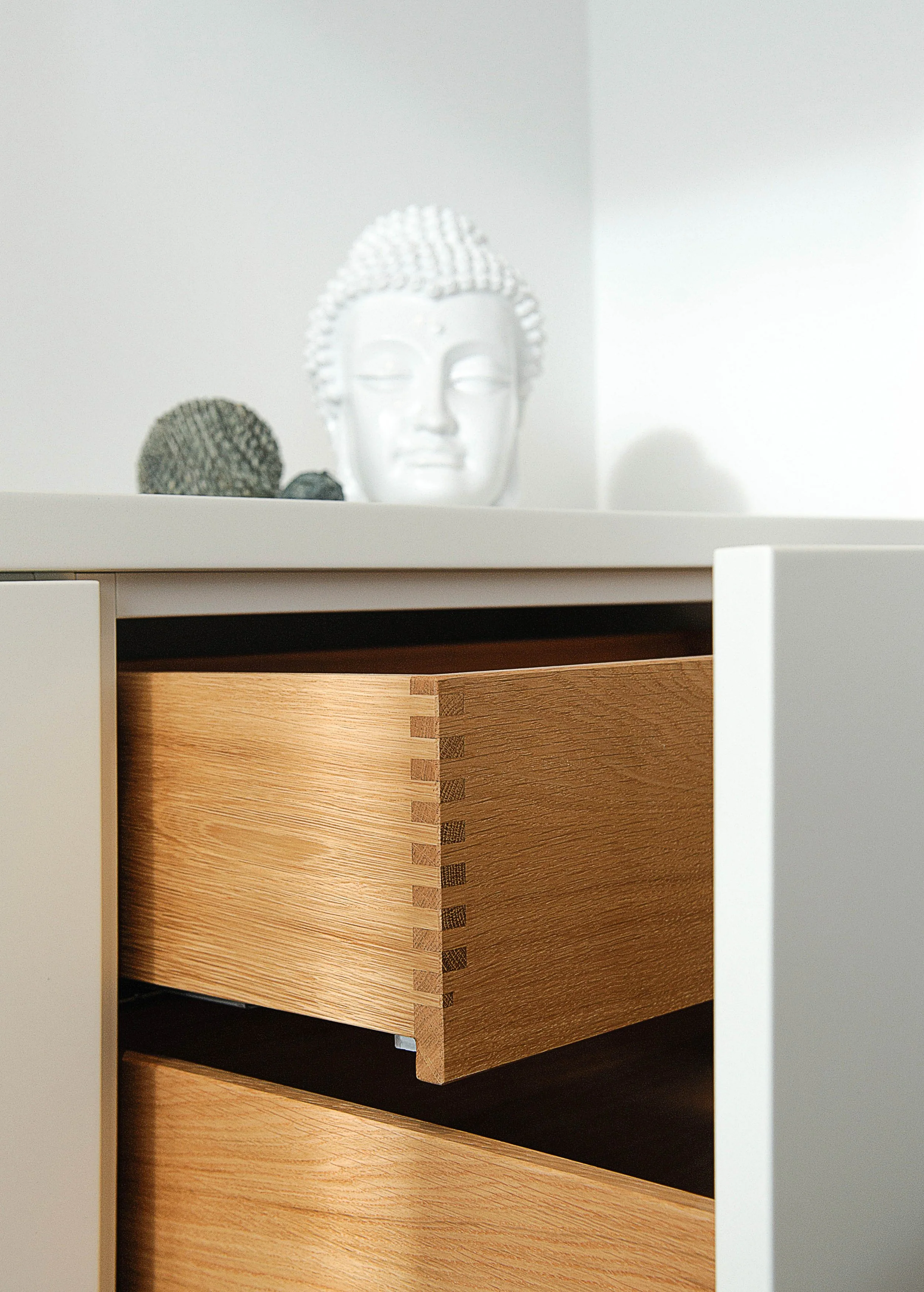 Nahaufnahme einer halb geöffneten Schublade aus Holz in einem weißen Möbelstück, mit einem dekorativen Buddha Kopf und Kakteen auf der Oberseite im Hintergrund.